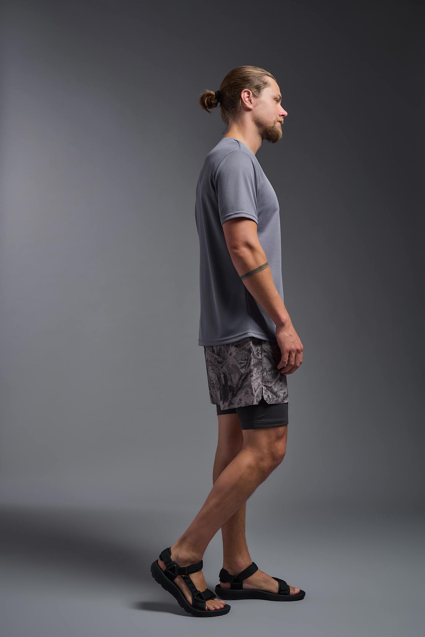 A blonde male model with a beard on a gray background, wearing the Men's Oxymesh™ Crewneck Tech Tee in the color steel gray from a full body side view. This shirt is made in the USA and is quick-drying and moisture-wicking. #color_steel-gray