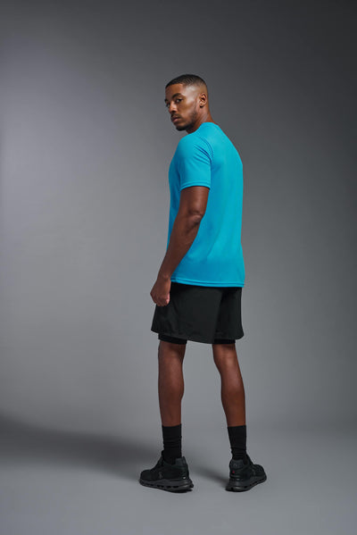 An African American male model with a beard on a gray background, wearing the Men's Oxymesh™ Crewneck Tech Tee in the color turquoise from a full body back view. This shirt is made in the USA and is quick-drying and moisture-wicking. #color_turquoise