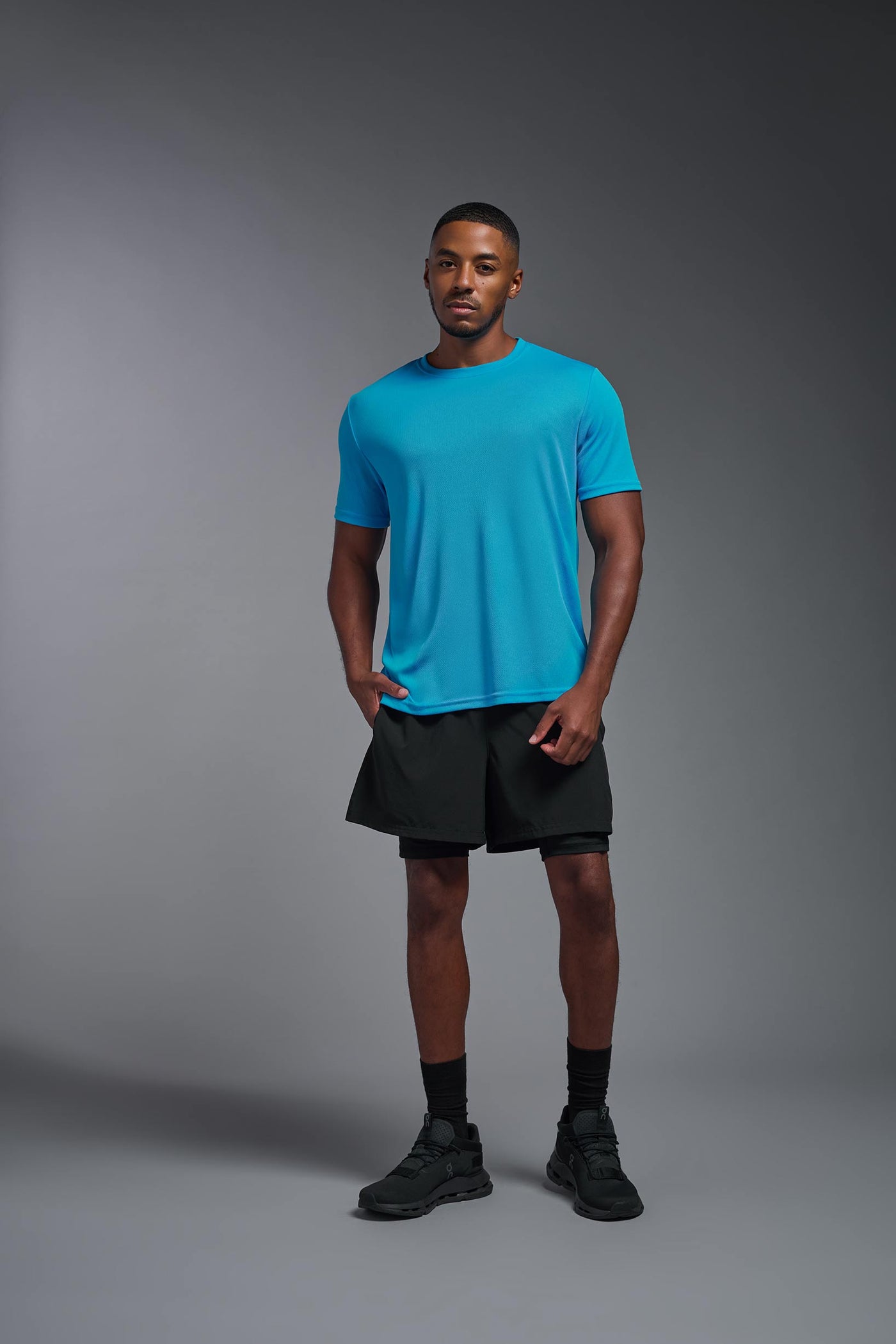An African American male model with a beard on a gray background, wearing the Men's Oxymesh™ Crewneck Tech Tee in the color turquoise from a full body front view. This shirt is made in the USA and is quick-drying and moisture-wicking. #color_turquoise