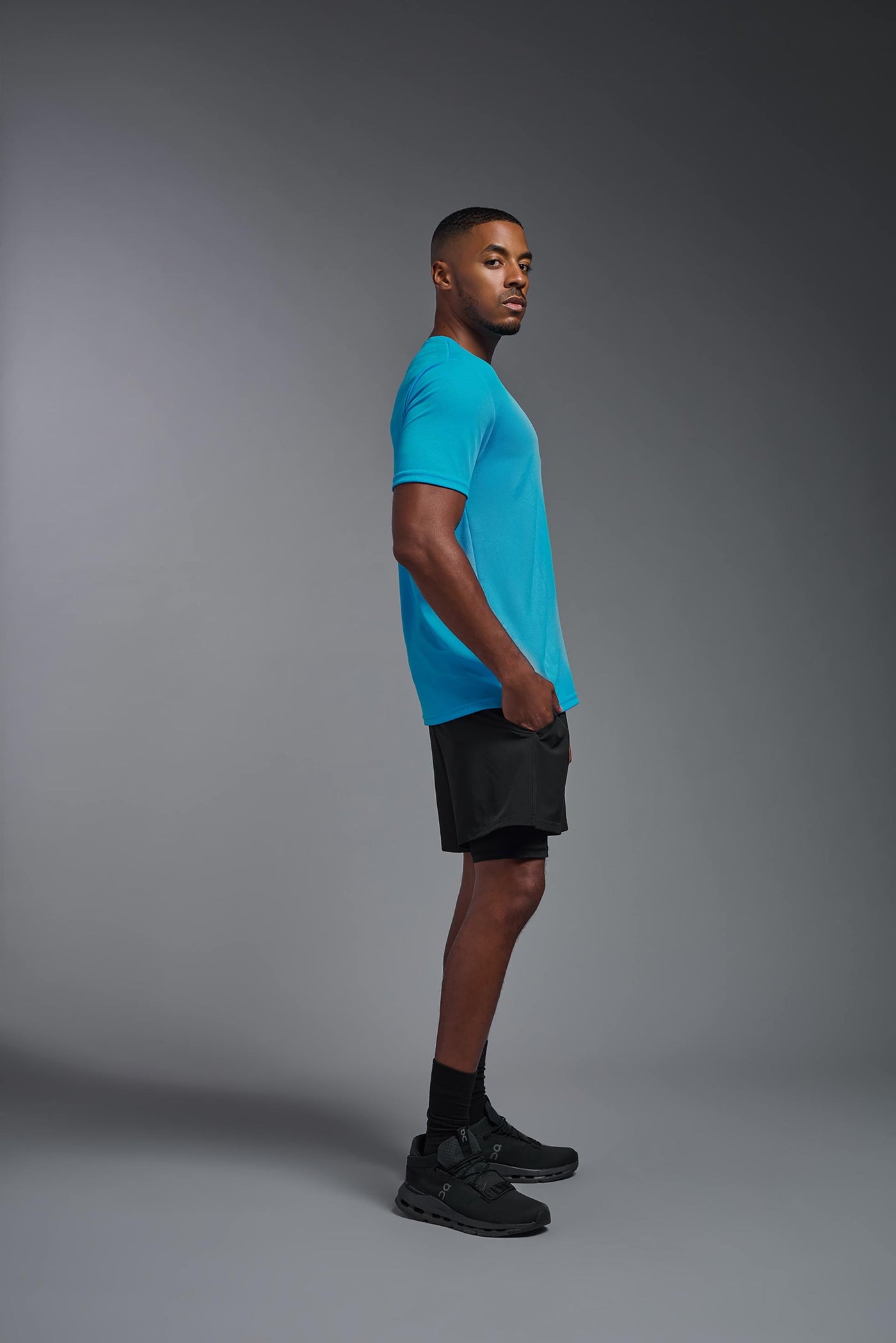 An African American male model with a beard on a gray background, wearing the Men's Oxymesh™ Crewneck Tech Tee in the color turquoise from a full body side view. This shirt is made in the USA and is quick-drying and moisture-wicking. #color_turquoise