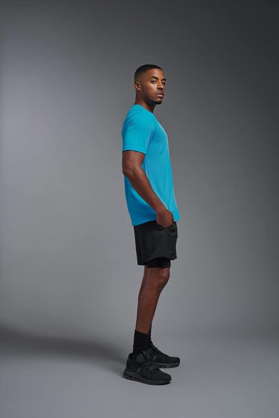 An African American male model with a beard on a gray background, wearing the Men's Oxymesh™ Crewneck Tech Tee in the color turquoise from a full body side view. This shirt is made in the USA and is quick-drying and moisture-wicking. #color_turquoise