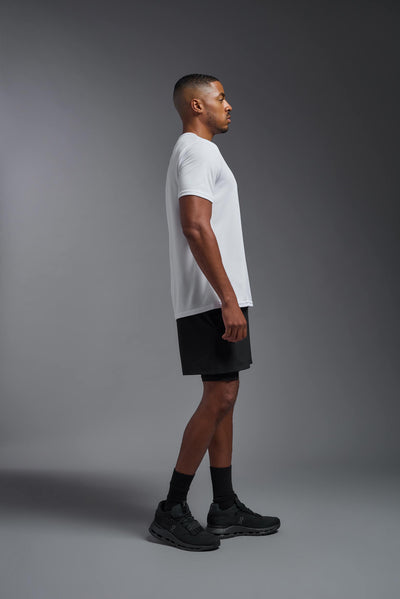 An African American male model with a beard on a gray background, wearing the Men's Oxymesh™ Crewneck Tech Tee in the color white from a full body side view. This shirt is made in the USA and is quick-drying and moisture-wicking. #color_white