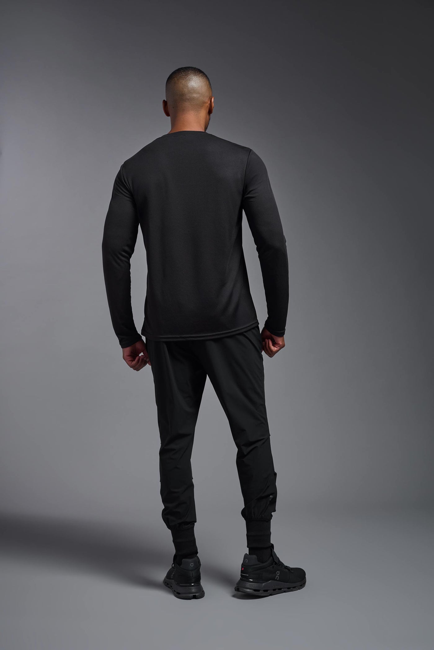 An African American male model with a beard wearing the men's Oxymesh Long Sleeve Tech Tee in the color black on a gray background, made out of 100% polyester, showing the relaxed fit from a full body back view. This shirt is breathable, quick-drying, moisture-wicking, and made in the USA. #color_black