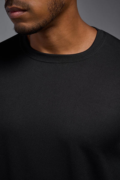 An African American male model with a beard wearing the men's Oxymesh Long Sleeve Tech Tee in the color black on a gray background, made out of 100% polyester, showing the relaxed fit from a closeup collar view. This shirt is breathable, quick-drying, moisture-wicking, and made in the USA. #color_black
