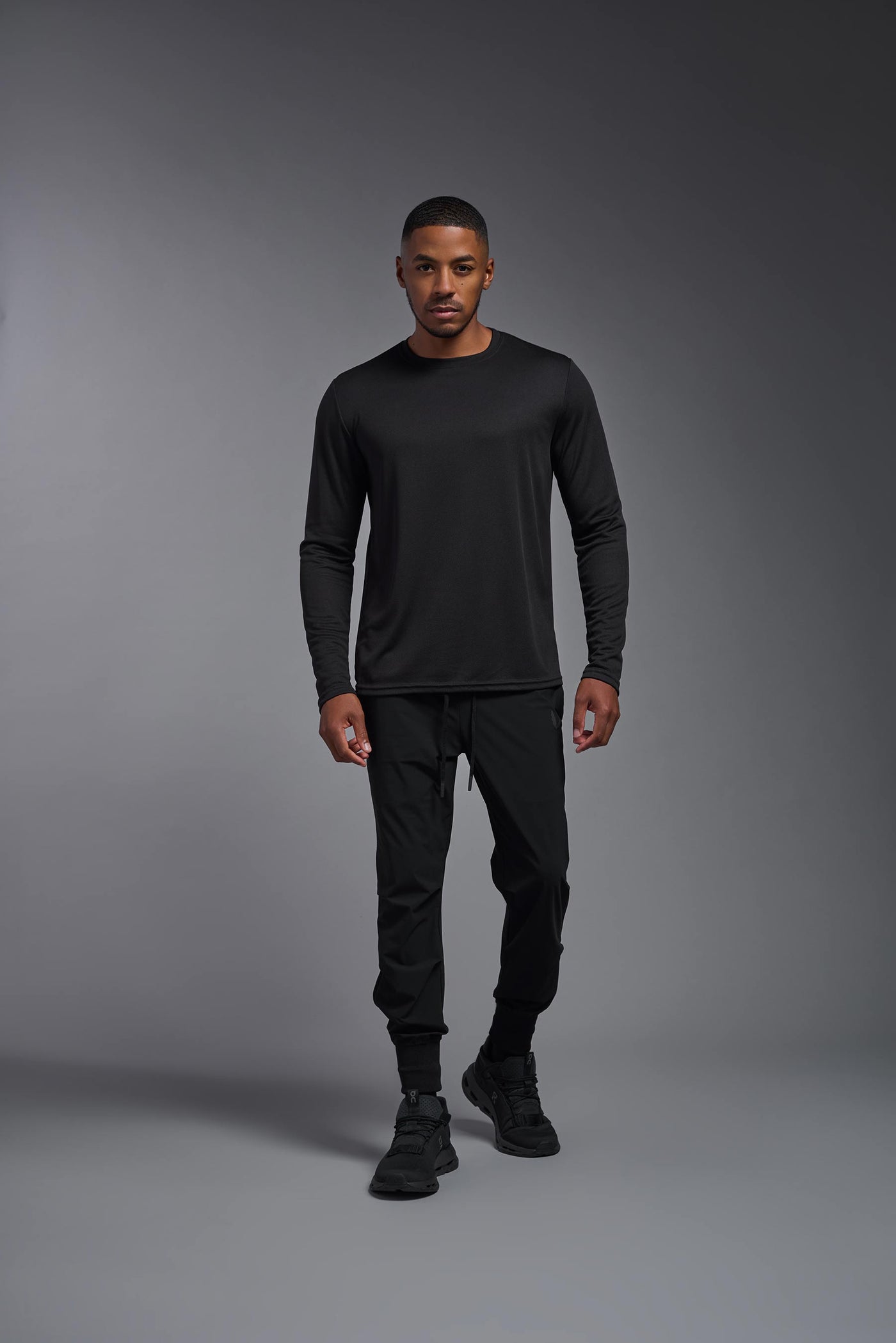 An African American male model with a beard wearing the men's Oxymesh Long Sleeve Tech Tee in the color black on a gray background, made out of 100% polyester, showing the relaxed fit from a full body front view. This shirt is breathable, quick-drying, moisture-wicking, and made in the USA. #color_black
