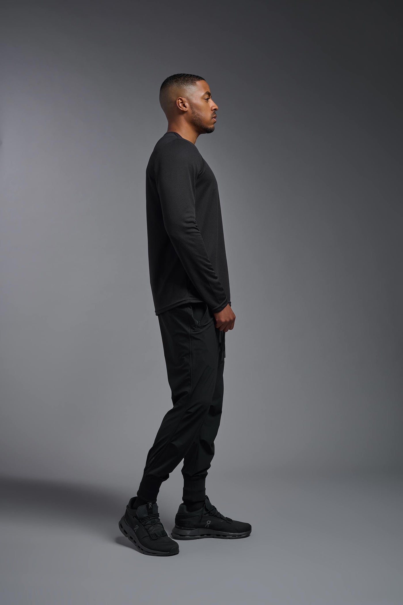 An African American male model with a beard wearing the men's Oxymesh Long Sleeve Tech Tee in the color black on a gray background, made out of 100% polyester, showing the relaxed fit from a full body side view. This shirt is breathable, quick-drying, moisture-wicking, and made in the USA. #color_black