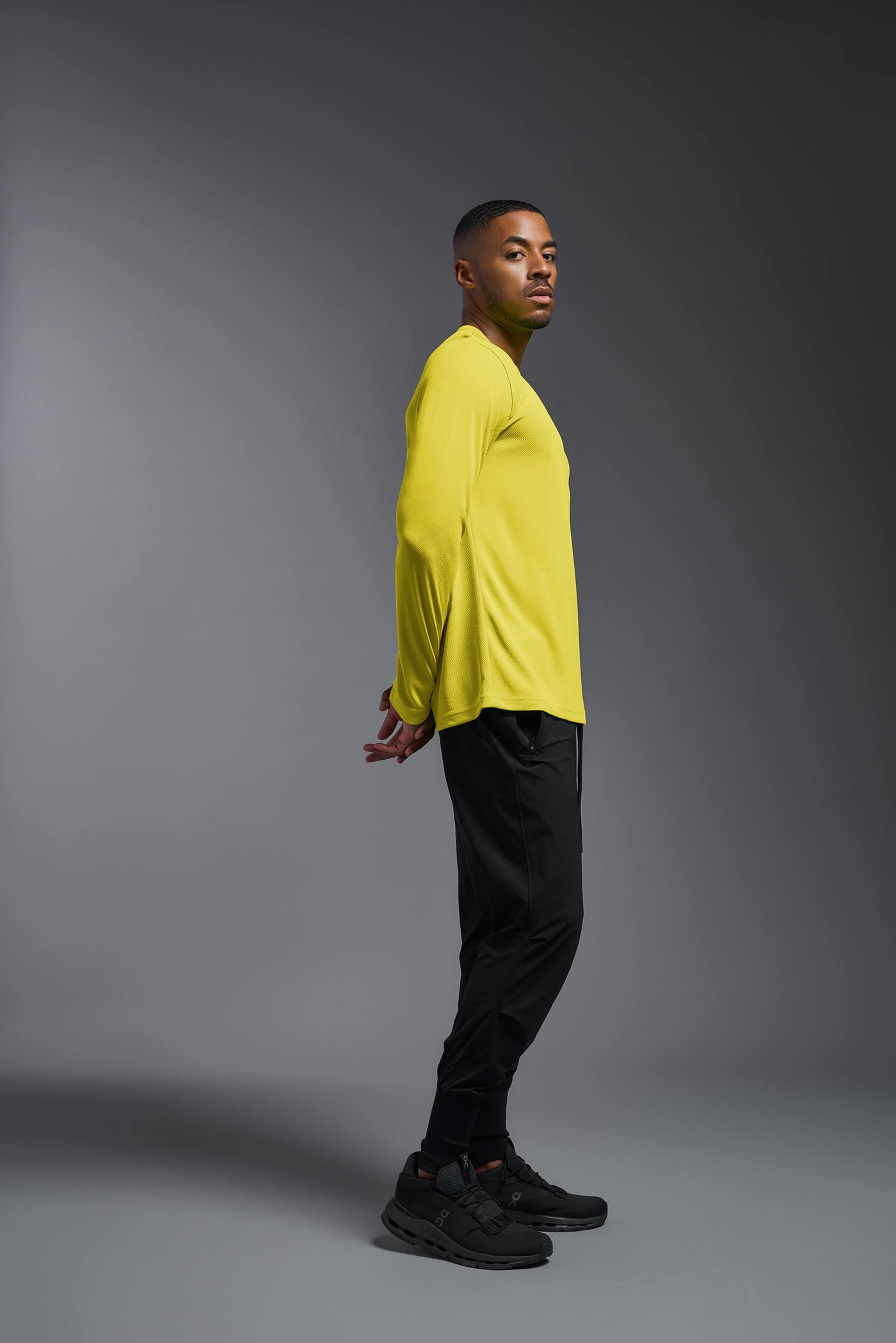 An African American male model with a beard wearing the men's Oxymesh Long Sleeve Tech Tee in the color bright yellow on a gray background, made out of 100% polyester, showing the relaxed fit from a full body side view. This shirt is breathable, quick-drying, moisture-wicking, and made in the USA. #color_bright-yellow