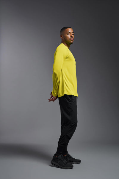 An African American male model with a beard wearing the men's Oxymesh Long Sleeve Tech Tee in the color bright yellow on a gray background, made out of 100% polyester, showing the relaxed fit from a full body side view. This shirt is breathable, quick-drying, moisture-wicking, and made in the USA. #color_bright-yellow