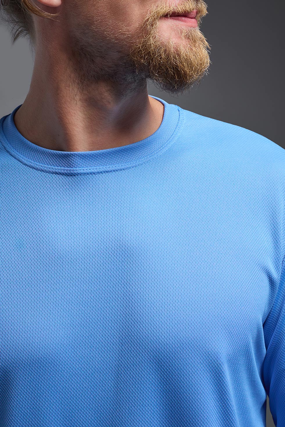 A blonde male model with a beard wearing the men's Oxymesh Long Sleeve Tech Tee in the color carolina blue on a gray background, made out of 100% polyester, showing the relaxed fit from a closeup collar front view. This shirt is breathable, quick-drying, moisture-wicking, and made in the USA. #color_carolina-blue