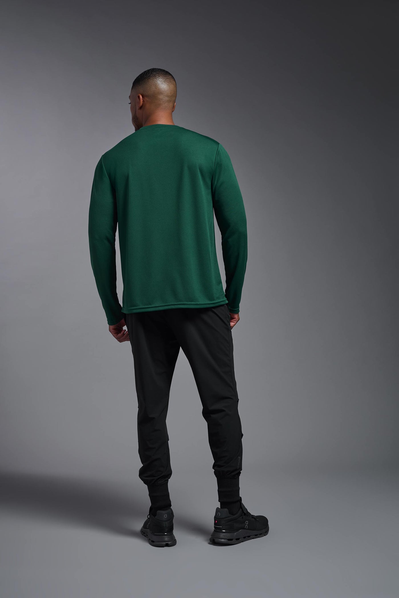 An African American male model with a beard wearing the men's Oxymesh Long Sleeve Tech Tee in the color forest green on a gray background, made out of 100% polyester, showing the relaxed fit from a full body back view. This shirt is breathable, quick-drying, moisture-wicking, and made in the USA. #color_forest-green