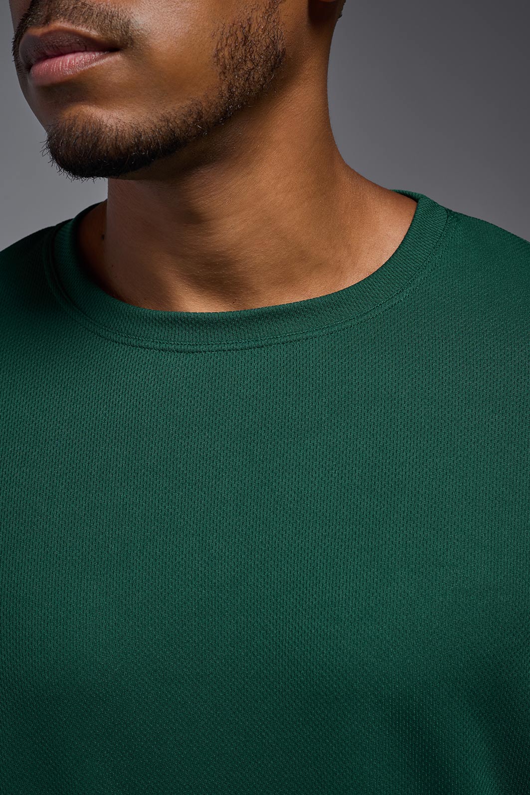 An African American male model with a beard wearing the men's Oxymesh Long Sleeve Tech Tee in the color forest green on a gray background, made out of 100% polyester, showing the relaxed fit from a closeup collar front view. This shirt is breathable, quick-drying, moisture-wicking, and made in the USA. #color_forest-green