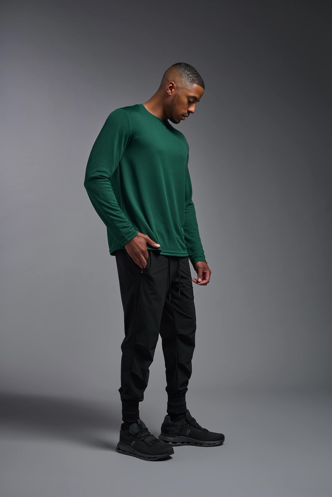 An African American male model with a beard wearing the men's Oxymesh Long Sleeve Tech Tee in the color forest green on a gray background, made out of 100% polyester, showing the relaxed fit from a full body side view. This shirt is breathable, quick-drying, moisture-wicking, and made in the USA. #color_forest-green