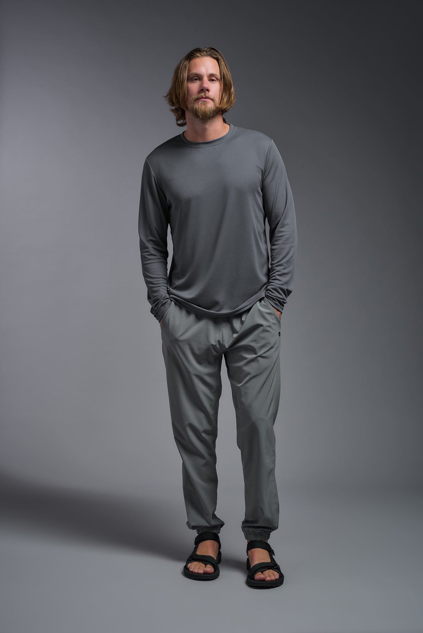 A blonde American male model with a beard wearing the men's Oxymesh Long Sleeve Tech Tee in the color graphite gray, made out of 100% polyester, showing the relaxed fit from a full body front view. This shirt is breathable, quick-drying, moisture-wicking, and made in the USA. #color_graphite-gray