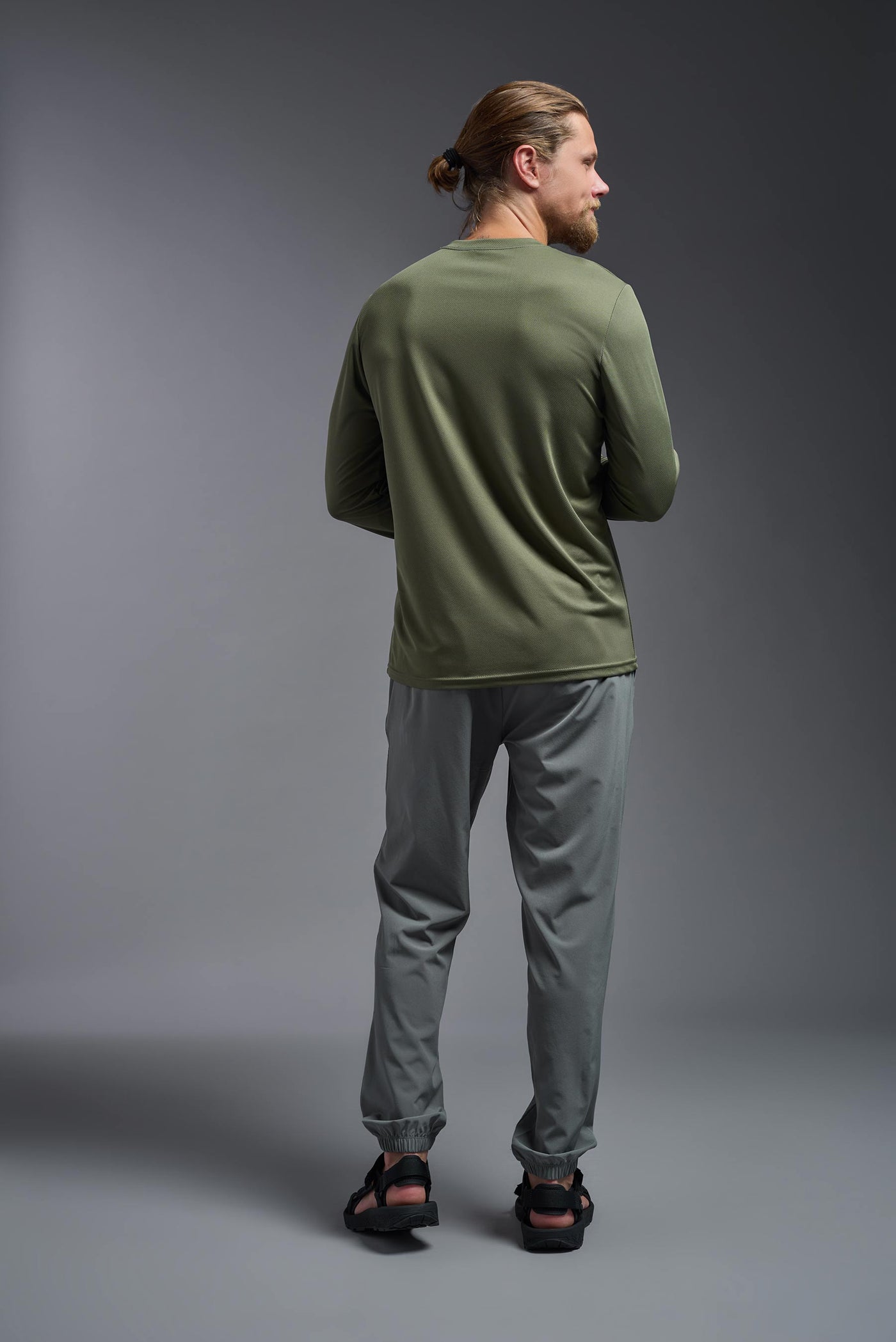 A blonde Caucasian male model with a beard wearing the men's Oxymesh Long Sleeve Tech Tee in the color military green on a gray background, made out of 100% polyester, showing the relaxed fit from a back view. This shirt is breathable, quick-drying, moisture-wicking, and made in the USA. #color_military-green