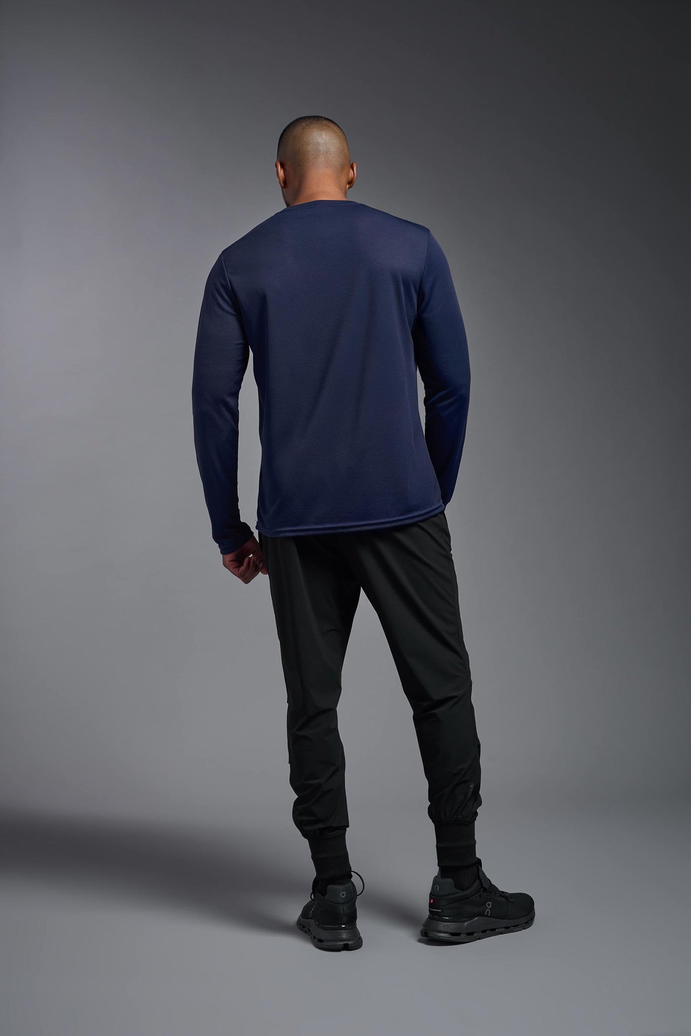 An African American male model with a beard wearing the men's Oxymesh Long Sleeve Tech Tee in the color navy on a gray background, made out of 100% polyester, showing the relaxed fit from a full body back view. This shirt is breathable, quick-drying, moisture-wicking, and made in the USA. #color_navy