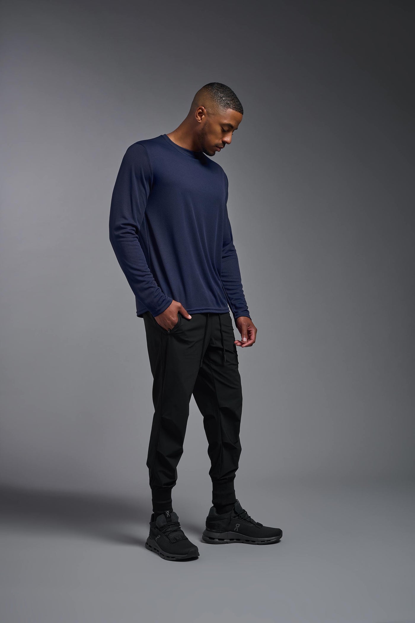 An African American male model with a beard wearing the men's Oxymesh Long Sleeve Tech Tee in the color navy on a gray background, made out of 100% polyester, showing the relaxed fit from a full body side view. This shirt is breathable, quick-drying, moisture-wicking, and made in the USA. #color_navy