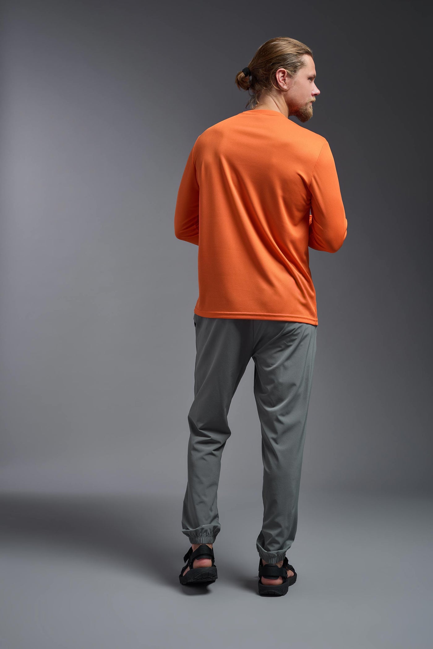 A blonde Caucasian model with a beard wearing the men's Oxymesh Long Sleeve Tech Tee in the color orange on a gray background, made out of 100% polyester, showing the relaxed fit from a full body back view. This shirt is breathable, quick-drying, moisture-wicking, and made in the USA. #color_orange