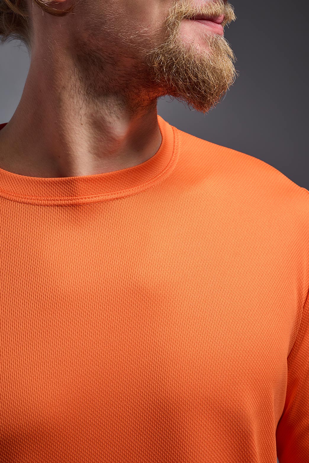 A blonde Caucasian model with a beard wearing the men's Oxymesh Long Sleeve Tech Tee in the color orange on a gray background, made out of 100% polyester, showing the relaxed fit from a closeup collar front view. This shirt is breathable, quick-drying, moisture-wicking, and made in the USA. #color_orange