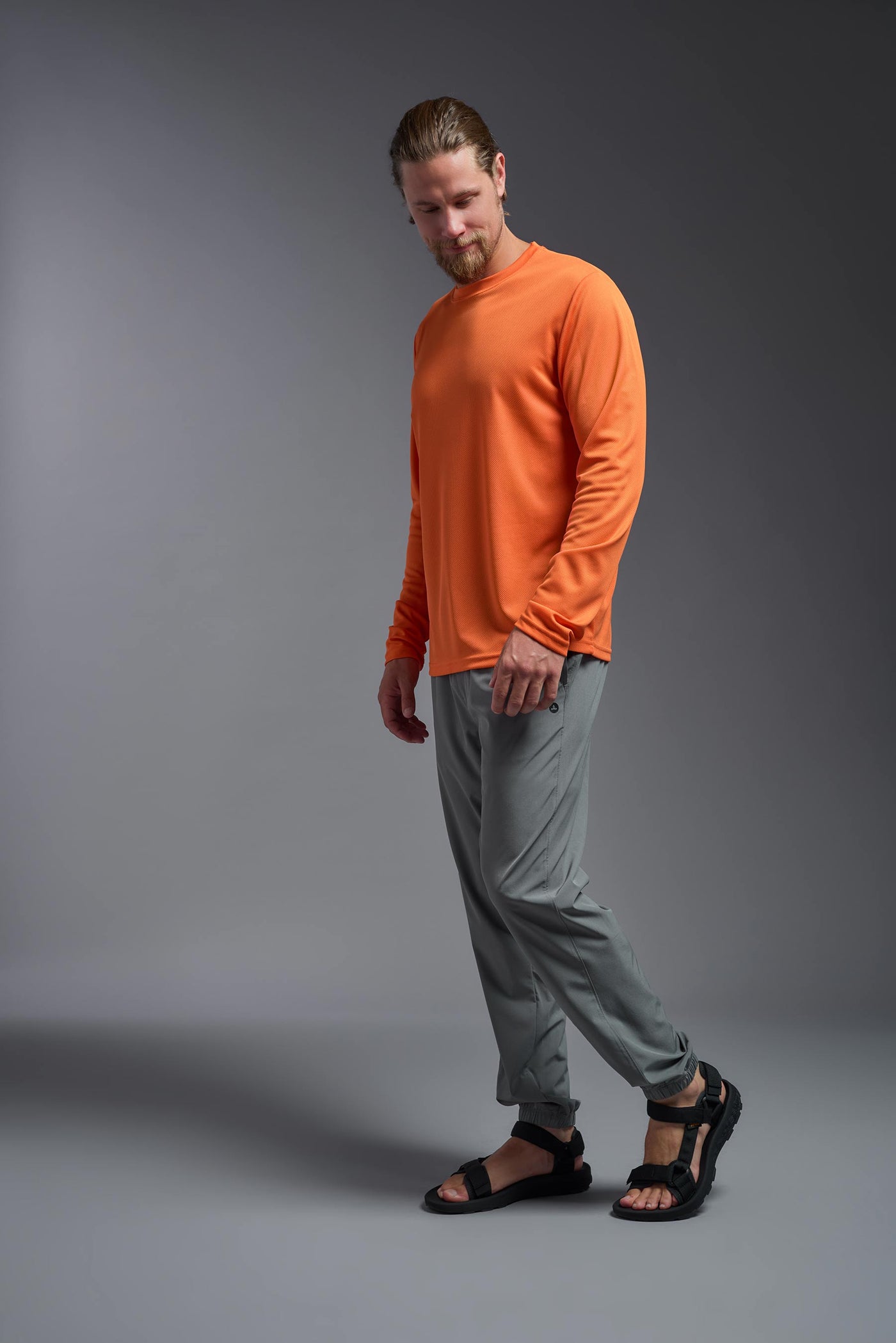 A blonde Caucasian model with a beard wearing the men's Oxymesh Long Sleeve Tech Tee in the color orange on a gray background, made out of 100% polyester, showing the relaxed fit from a full body side view. This shirt is breathable, quick-drying, moisture-wicking, and made in the USA. #color_orange