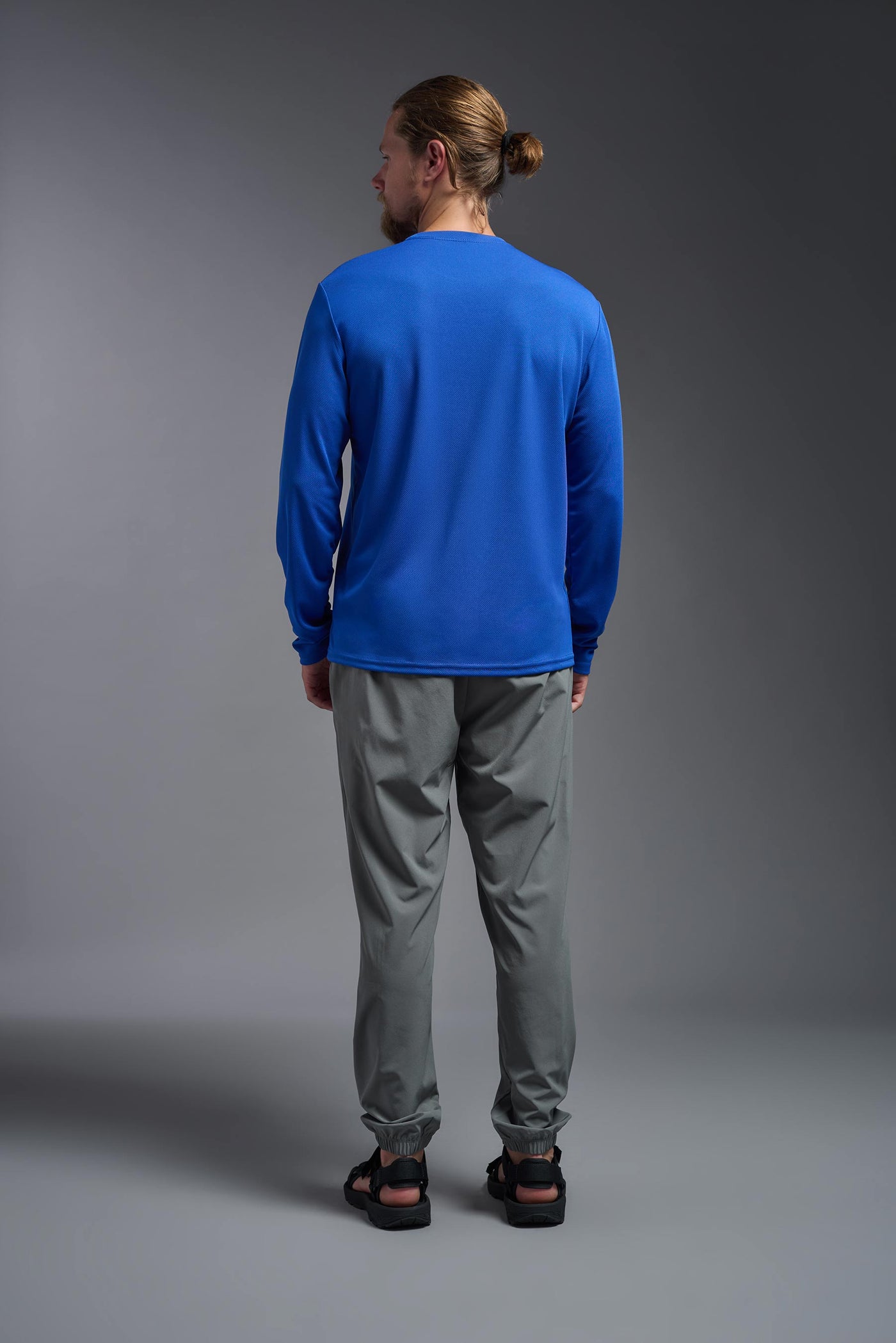 A blonde male model with a beard wearing the men's Oxymesh Long Sleeve Tech Tee in the color royal blue on a gray background, made out of 100% polyester, showing the relaxed fit from a full body back view. This shirt is breathable, quick-drying, moisture-wicking, and made in the USA. #color_royal-blue