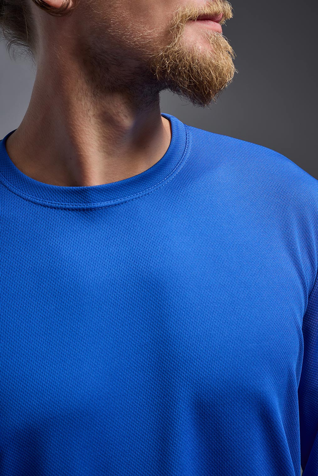 A blonde male model with a beard wearing the men's Oxymesh Long Sleeve Tech Tee in the color royal blue on a gray background, made out of 100% polyester, showing the relaxed fit from a closeup collar front view. This shirt is breathable, quick-drying, moisture-wicking, and made in the USA. #color_royal-blue