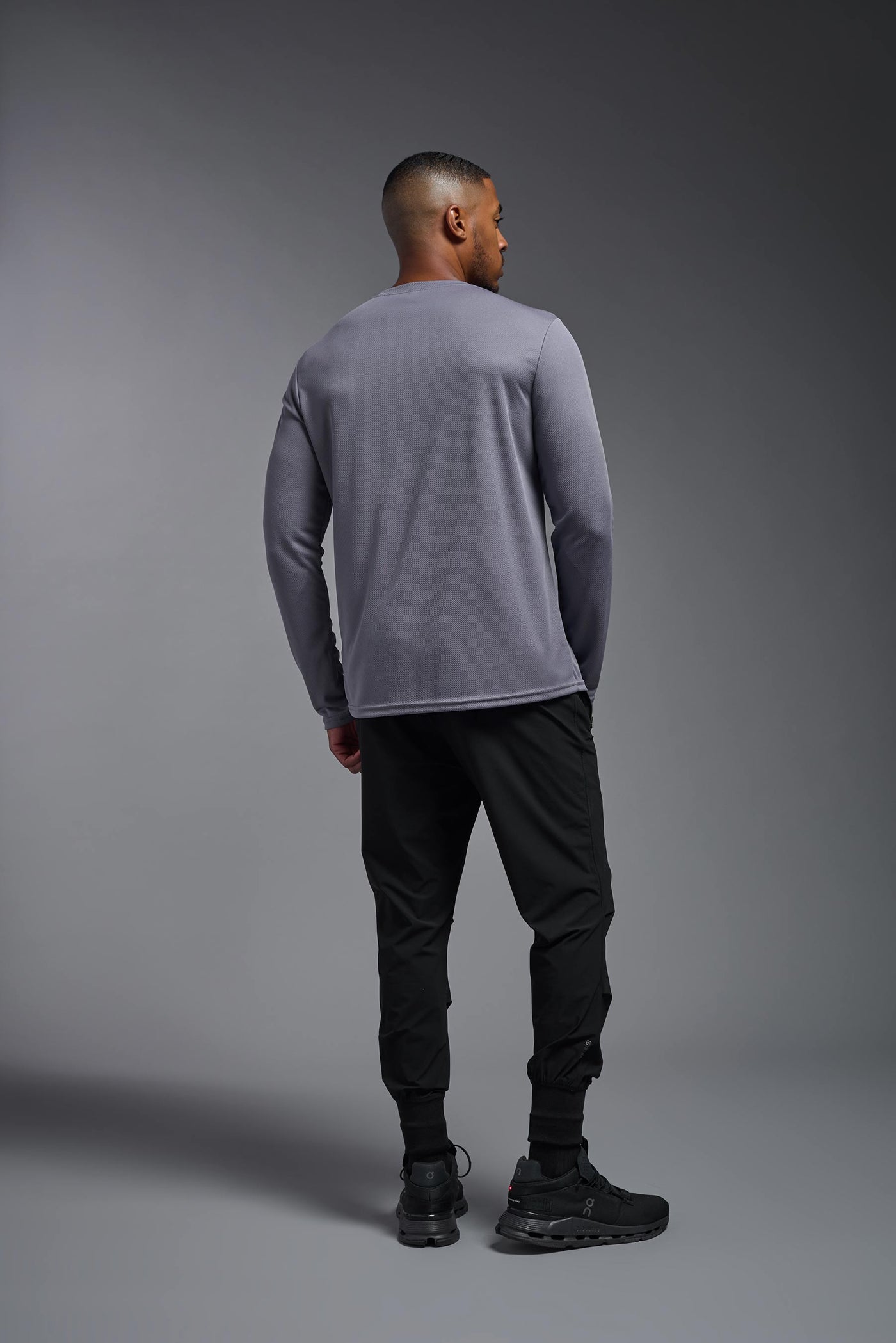 An African American male model with a beard wearing the men's Oxymesh Long Sleeve Tech Tee in the color steel gray on a gray background, made out of 100% polyester, showing the relaxed fit from a full body back view. This shirt is breathable, quick-drying, moisture-wicking, and made in the USA. #color_steel-gray