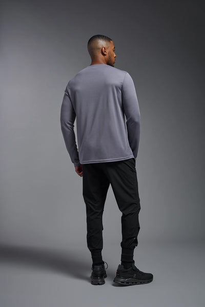 An African American male model with a beard wearing the men's Oxymesh Long Sleeve Tech Tee in the color steel gray on a gray background, made out of 100% polyester, showing the relaxed fit from a full body back view. This shirt is breathable, quick-drying, moisture-wicking, and made in the USA. #color_steel-gray