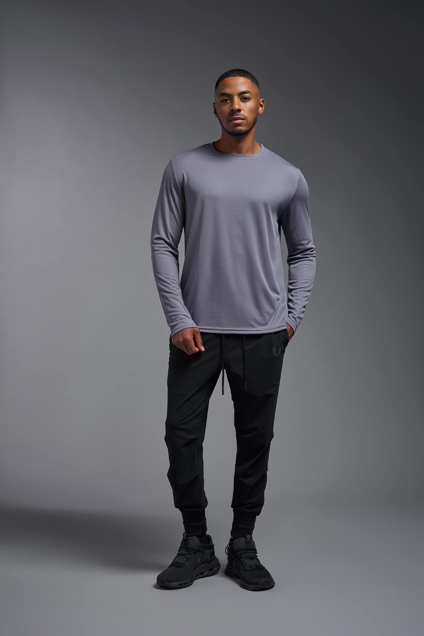 An African American male model with a beard wearing the men's Oxymesh Long Sleeve Tech Tee in the color steel gray on a gray background, made out of 100% polyester, showing the relaxed fit from a full body front view. This shirt is breathable, quick-drying, moisture-wicking, and made in the USA. #color_steel-gray