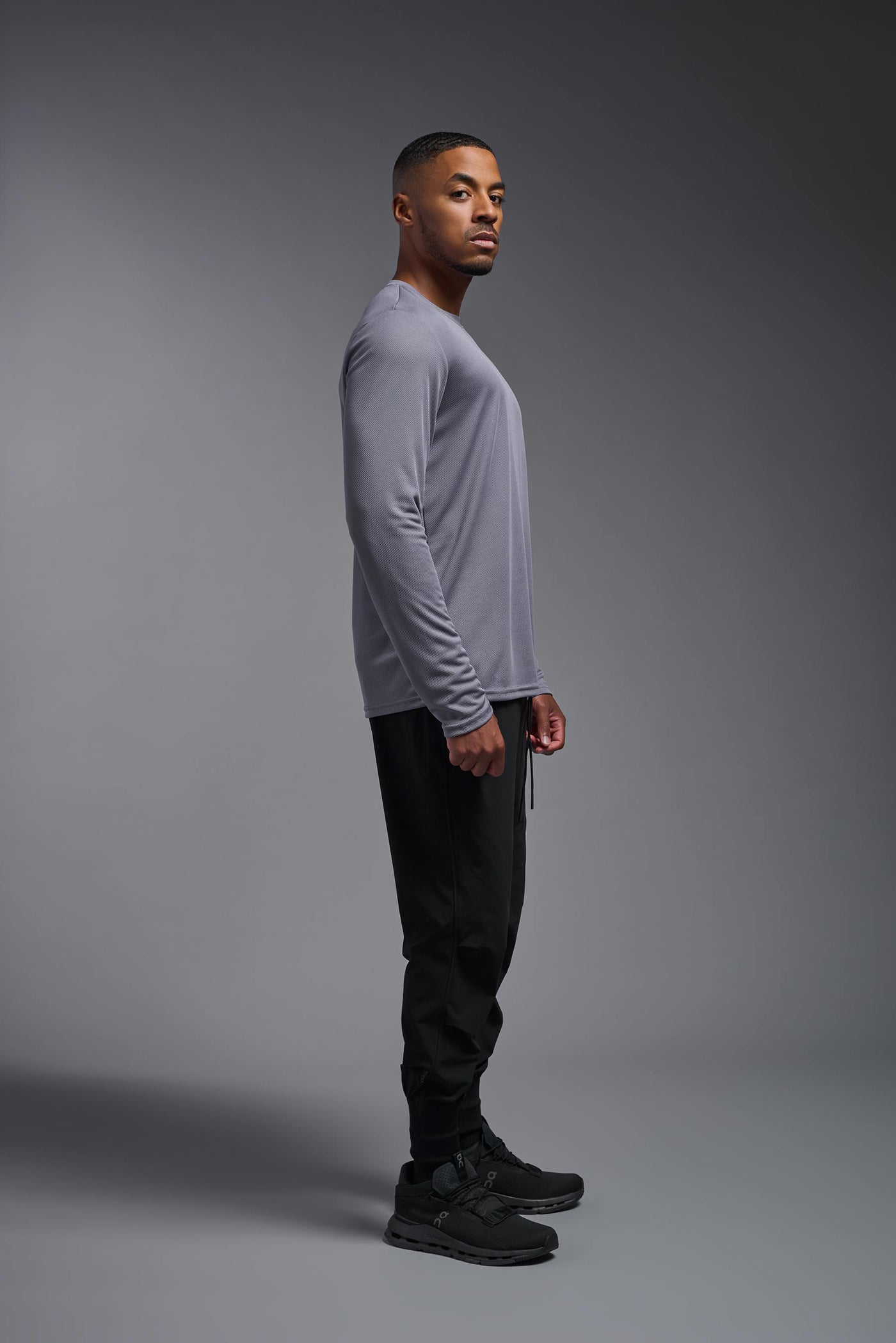 An African American male model with a beard wearing the men's Oxymesh Long Sleeve Tech Tee in the color steel gray on a gray background, made out of 100% polyester, showing the relaxed fit from a full body side view. This shirt is breathable, quick-drying, moisture-wicking, and made in the USA. #color_steel-gray