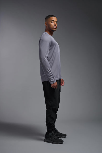 An African American male model with a beard wearing the men's Oxymesh Long Sleeve Tech Tee in the color steel gray on a gray background, made out of 100% polyester, showing the relaxed fit from a full body side view. This shirt is breathable, quick-drying, moisture-wicking, and made in the USA. #color_steel-gray