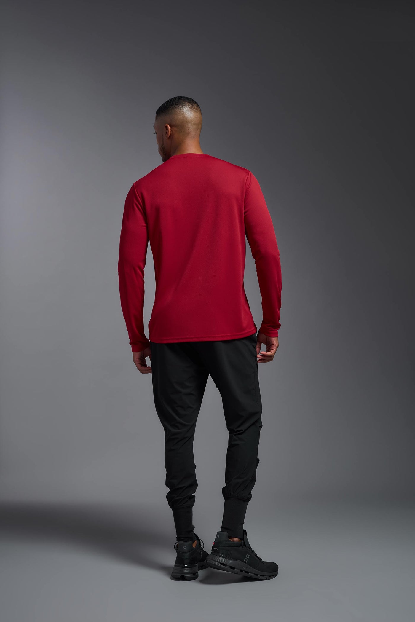An African American male model with a beard wearing the men's Oxymesh Long Sleeve Tech Tee in the color true red on a gray background, made out of 100% polyester, showing the relaxed fit from a full body back view. This shirt is breathable, quick-drying, moisture-wicking, and made in the USA. #color_true-red