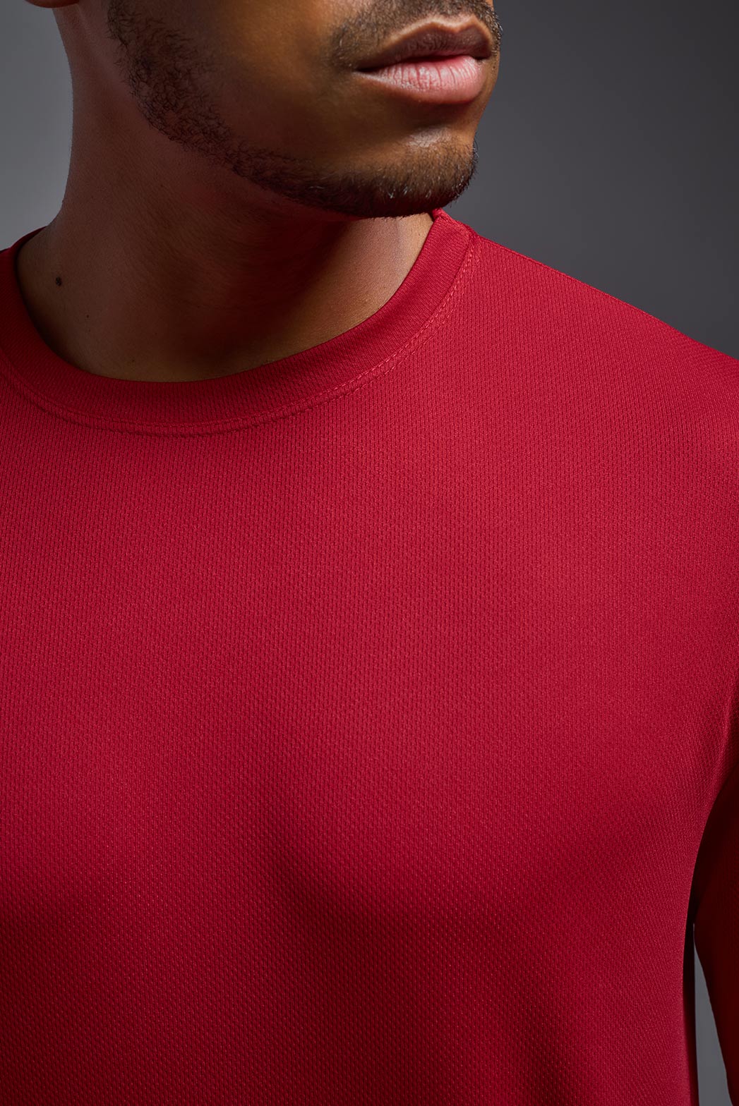 An African American male model with a beard wearing the men's Oxymesh Long Sleeve Tech Tee in the color true red on a gray background, made out of 100% polyester, showing the relaxed fit from a closeup collar front view. This shirt is breathable, quick-drying, moisture-wicking, and made in the USA. #color_true-red
