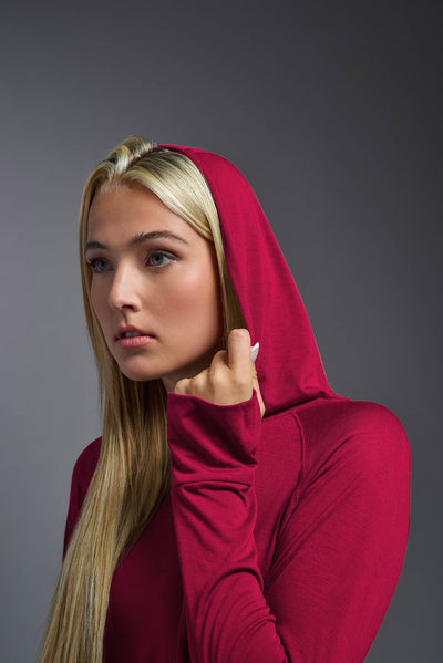 A blonde female model with long straight hair on a gray background, wearing the Women's Siro Curvy Hoodie Shirt in the color scarlet from a closeup up front view featuring the hood up. This hoodie shirt is made in the USA and is quick-drying and moisture-wicking. #color_scarlet
