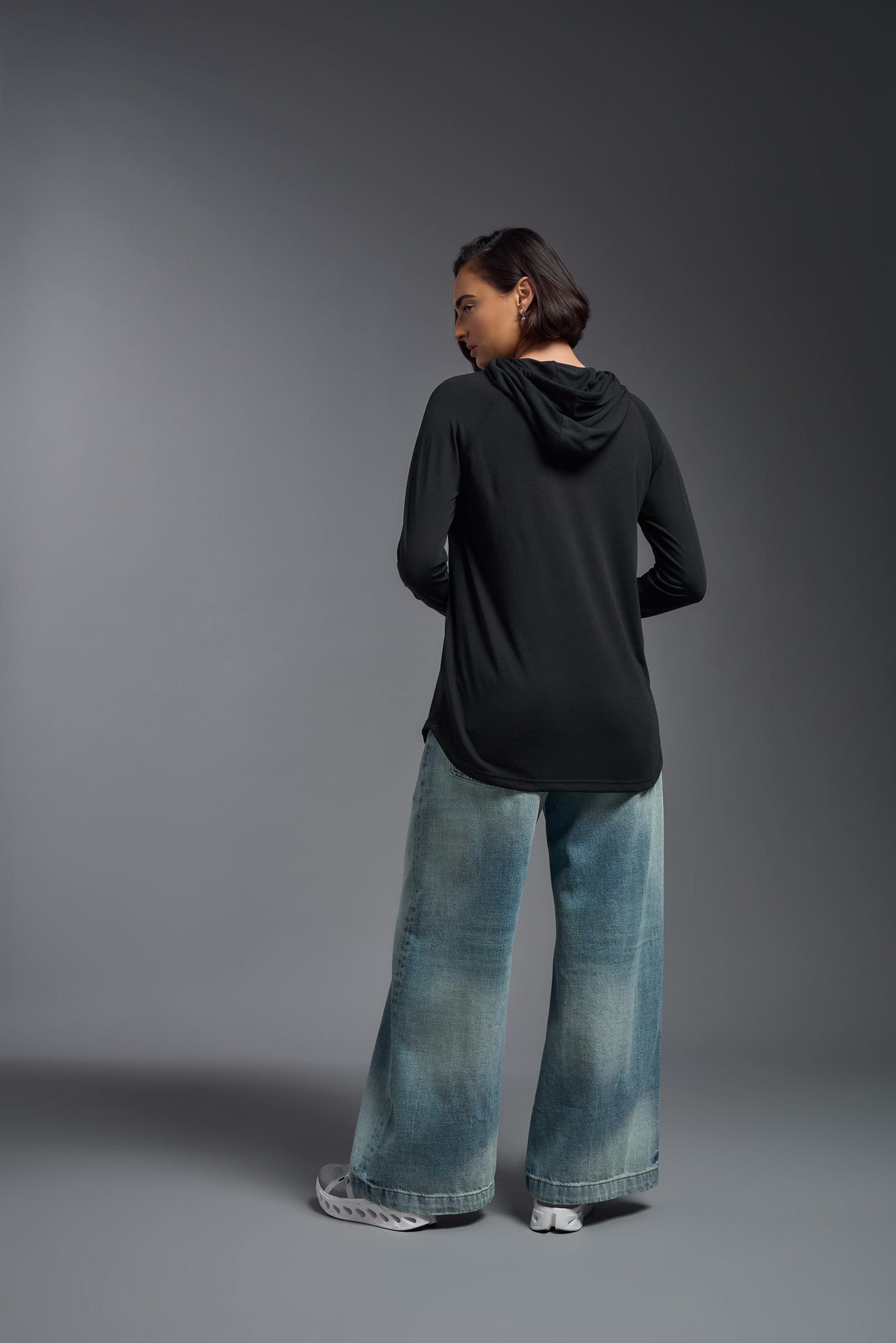A female model with short brown hair on a gray background, wearing the Women's Siro Curvy Hoodie Shirt in the color black from a full body back view. This hoodie shirt is made in the USA and is quick-drying and moisture-wicking. #color_black