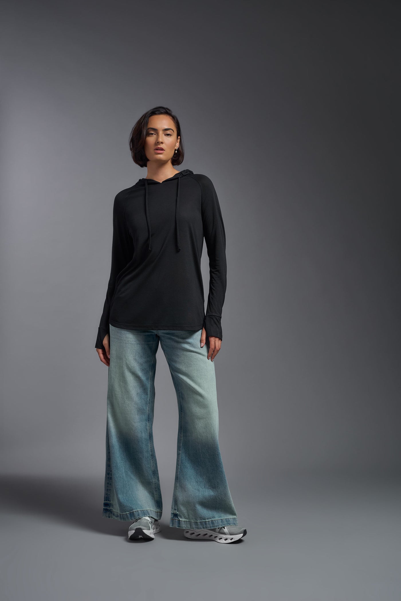 A female model with short brown hair on a gray background, wearing the Women's Siro Curvy Hoodie Shirt in the color black from a full body front view. This hoodie shirt is made in the USA and is quick-drying and moisture-wicking. #color_black
