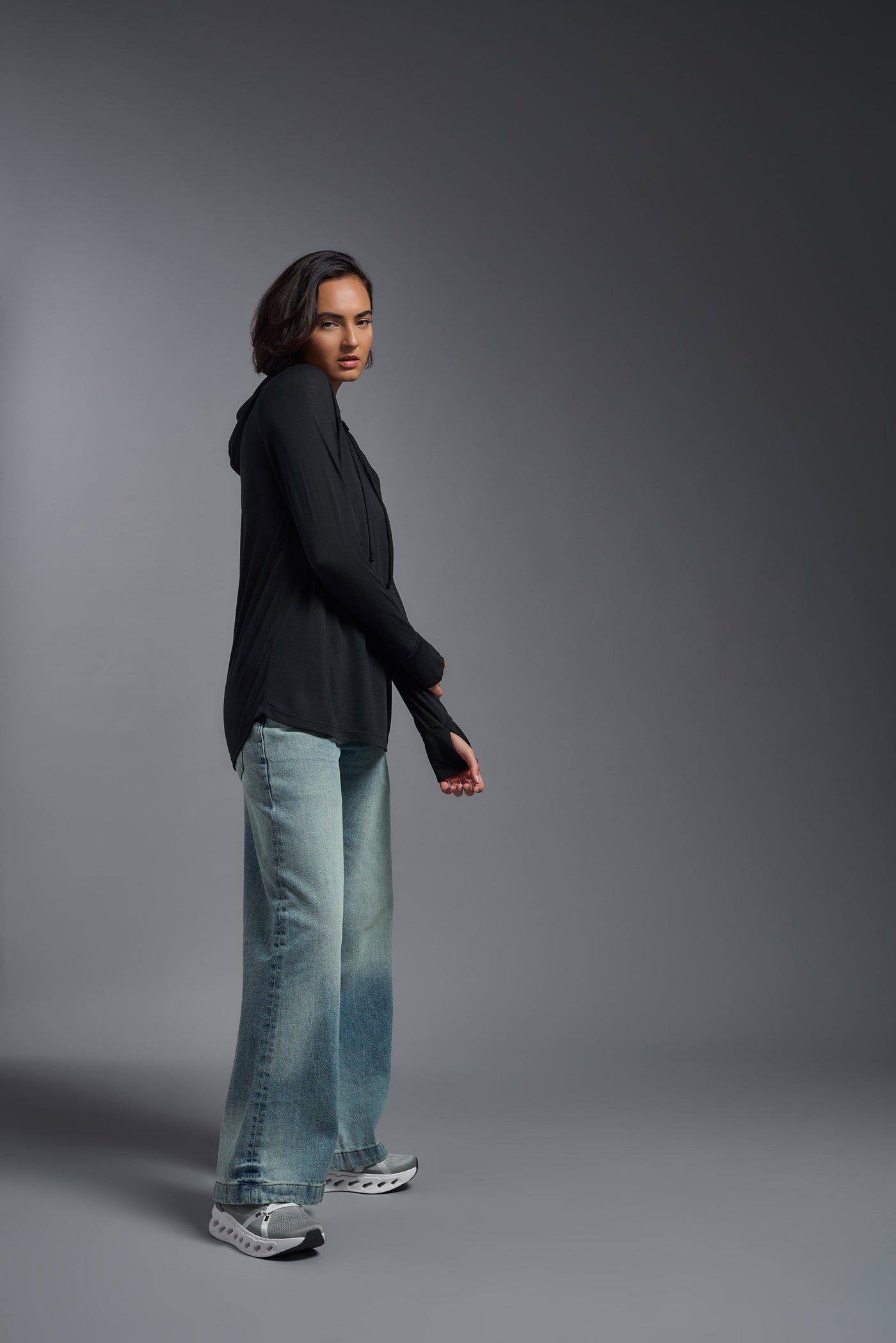 A female model with short brown hair on a gray background, wearing the Women's Siro Curvy Hoodie Shirt in the color black from a full body side view. This hoodie shirt is made in the USA and is quick-drying and moisture-wicking. #color_black