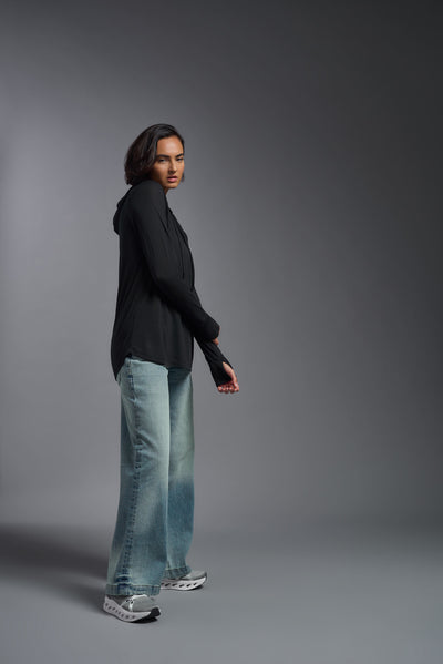A female model with short brown hair on a gray background, wearing the Women's Siro Curvy Hoodie Shirt in the color black from a full body side view. This hoodie shirt is made in the USA and is quick-drying and moisture-wicking. #color_black