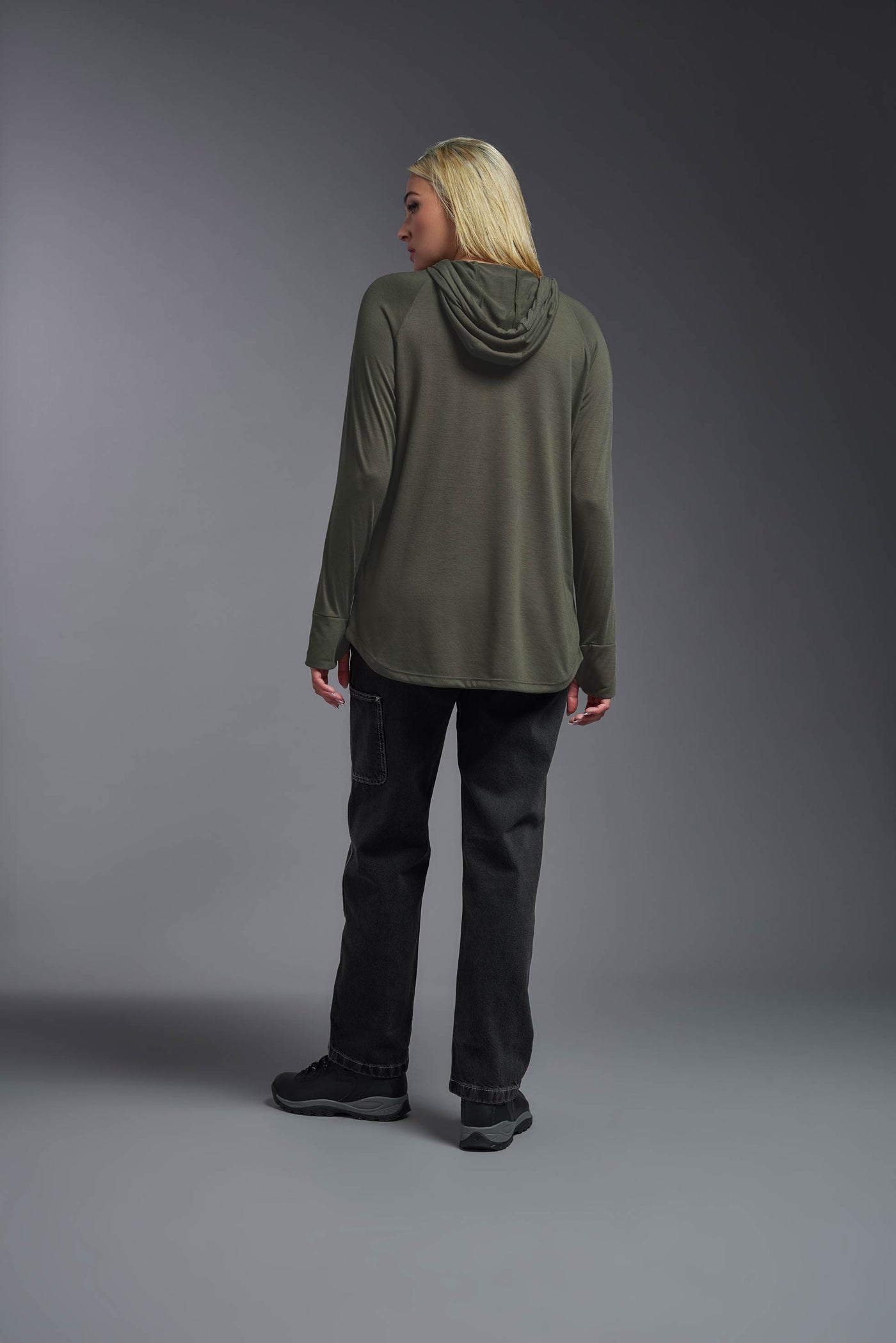 A blonde female model with long straight hair on a gray background, wearing the Women's Siro Curvy Hoodie Shirt in the color olive green from a full body back view. This hoodie shirt is made in the USA and is quick-drying and moisture-wicking. #color_olive