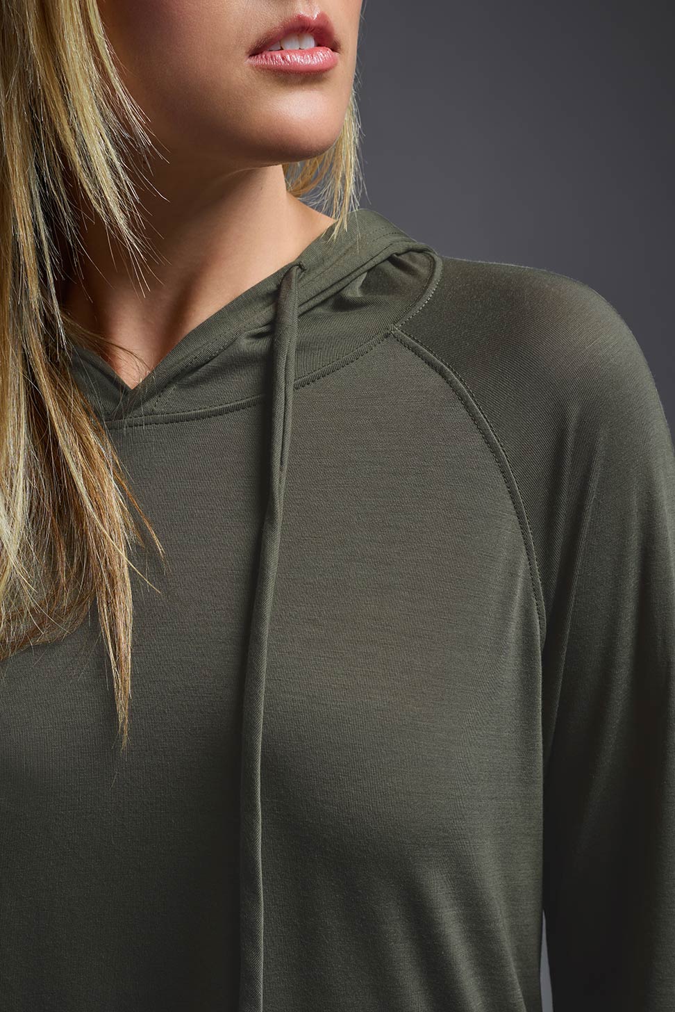 A blonde female model with long straight hair on a gray background, wearing the Women's Siro Curvy Hoodie Shirt in the color olive green from a closeup collar detail view. This hoodie shirt is made in the USA and is quick-drying and moisture-wicking. #color_olive