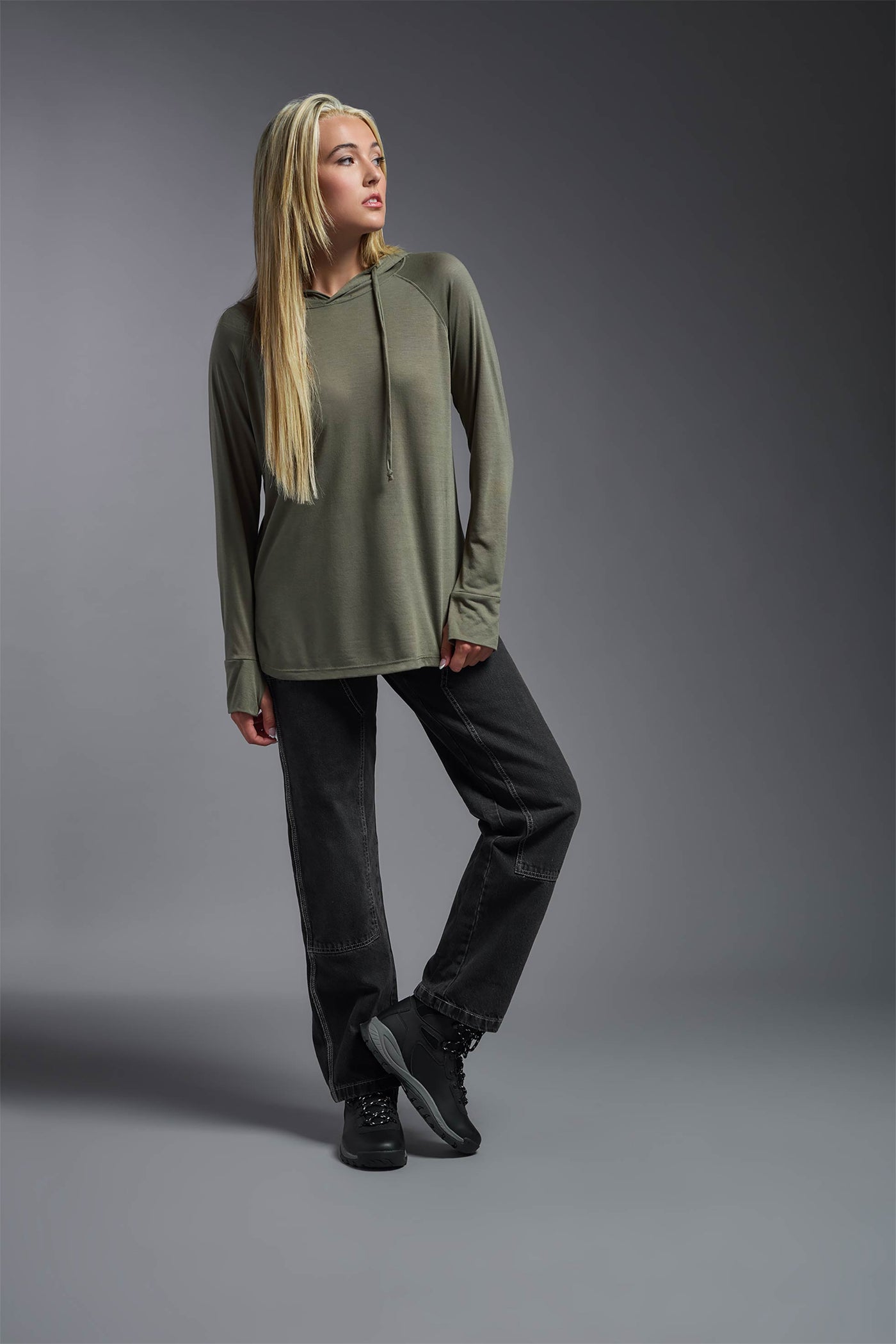 A blonde female model with long straight hair on a gray background, wearing the Women's Siro Curvy Hoodie Shirt in the color olive green from a full body front view. This hoodie shirt is made in the USA and is quick-drying and moisture-wicking. #color_olive