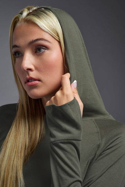 A blonde female model with long straight hair on a gray background, wearing the Women's Siro Curvy Hoodie Shirt in the color olive green from a full body front hooded view. This hoodie shirt is made in the USA and is quick-drying and moisture-wicking. #color_olive