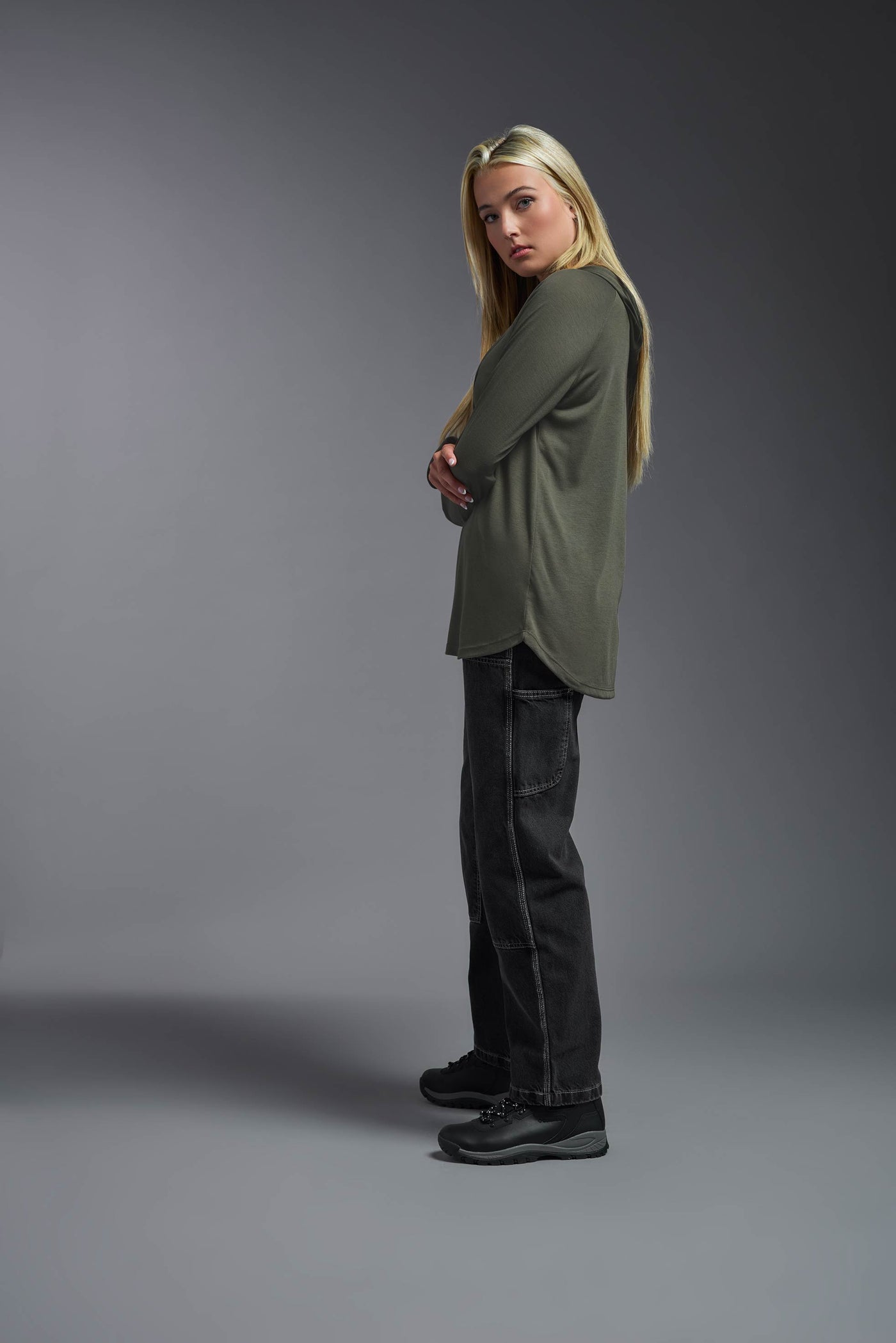 A blonde female model with long straight hair on a gray background, wearing the Women's Siro Curvy Hoodie Shirt in the color olive green from a full body side view. This hoodie shirt is made in the USA and is quick-drying and moisture-wicking. #color_olive