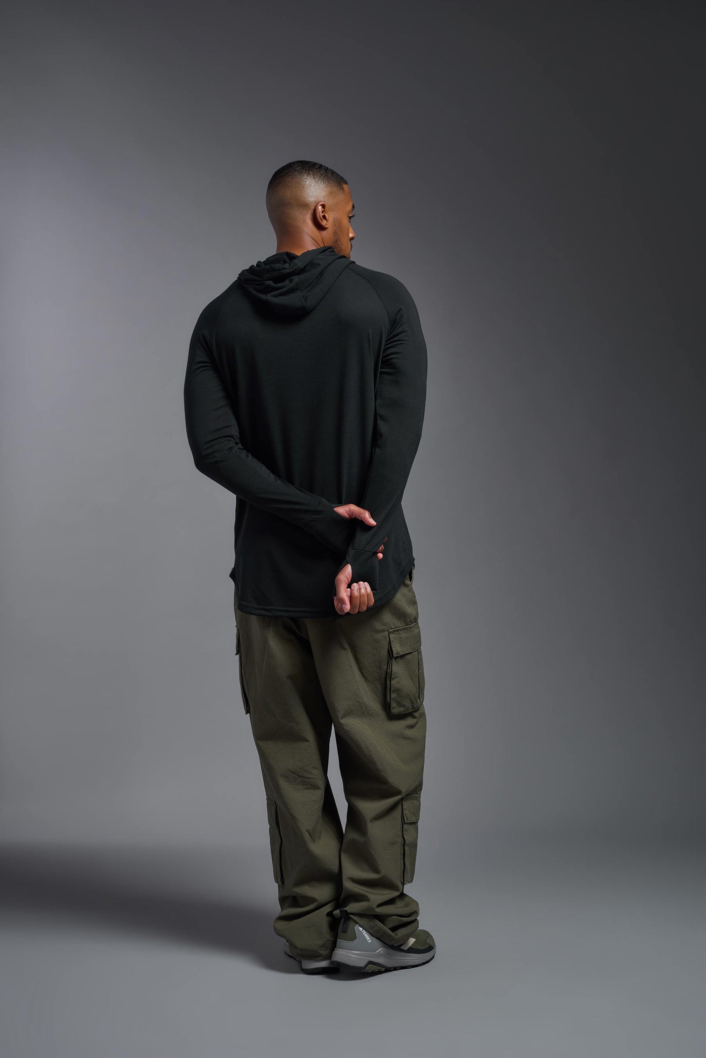 An African American, male model with a beard wearing the men's Siro Curvy Hoodie Shirt in the color black back, made out of 100% polyester, showing the relaxed fit from a back view. This shirt features a soft cotton feel, quick-dries, wicks moisture and is made in the USA. #color_black
