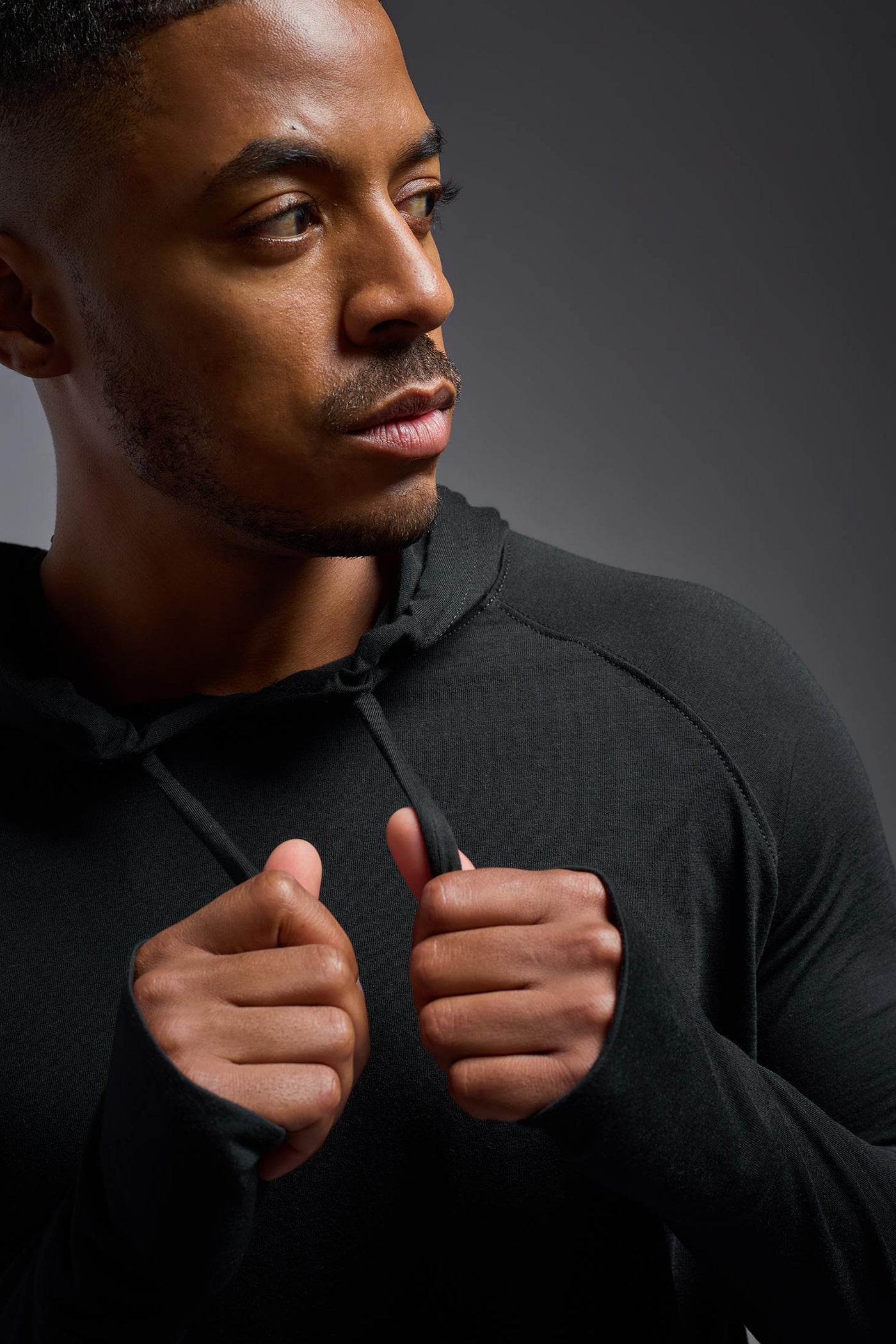An African American, male model with a beard wearing the men's Siro Curvy Hoodie Shirt in the color black, made out of 100% polyester, showing the relaxed fit from a front detail collar view. This shirt features a soft cotton feel, quick-dries, wicks moisture and is made in the USA. #color_black