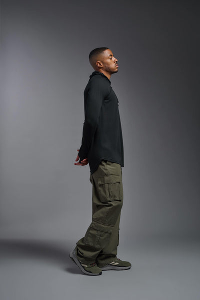 An African American, male model with a beard wearing the men's Siro Curvy Hoodie Shirt in the color black, made out of 100% polyester, showing the relaxed fit from a side view. This shirt features a soft cotton feel, quick-dries, wicks moisture and is made in the USA. #color_black