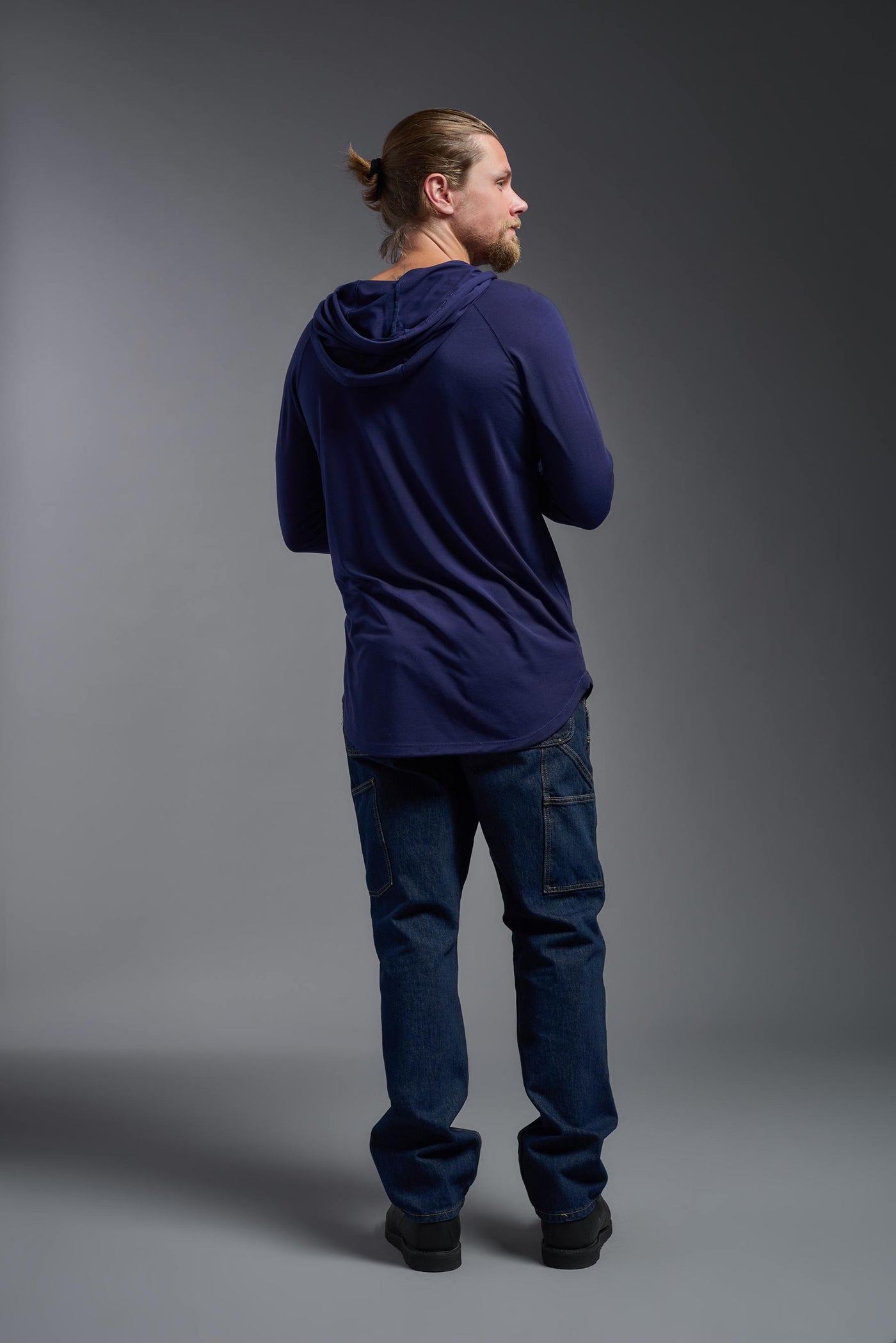 A blonde male model with a beard wearing the men's Siro Curvy Hoodie Shirt in the color navy, made out of 100% polyester, showing the relaxed fit from a back view. This hoodie shirt features a soft cotton feel, quick-dries, wicks moisture and is made in the USA. #color_navy