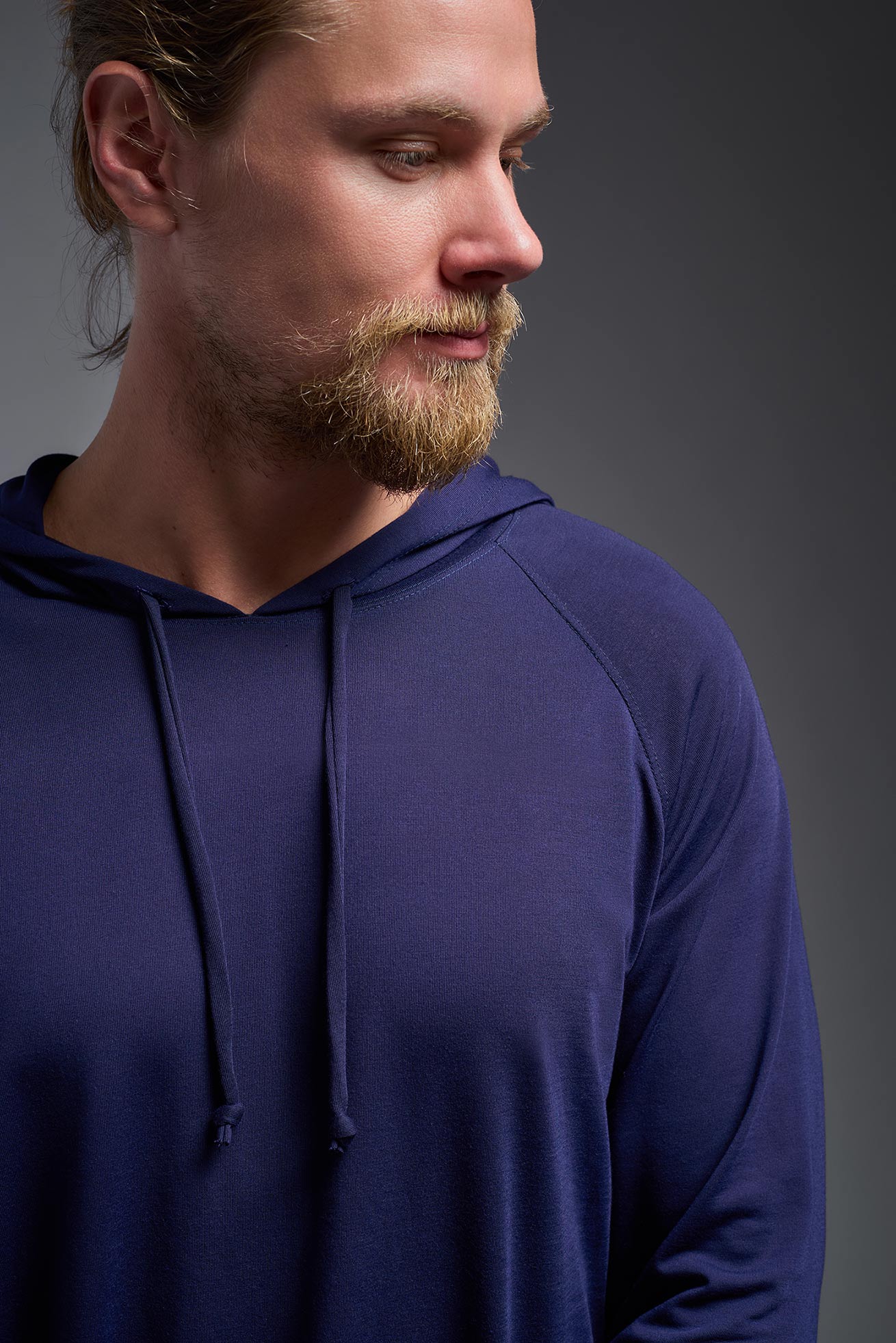 A blonde male model with a beard wearing the men's Siro Curvy Hoodie Shirt in the color navy, made out of 100% polyester, showing the relaxed fit from a front collar closeup view. This hoodie shirt features a soft cotton feel, quick-dries, wicks moisture and is made in the USA. #color_navy