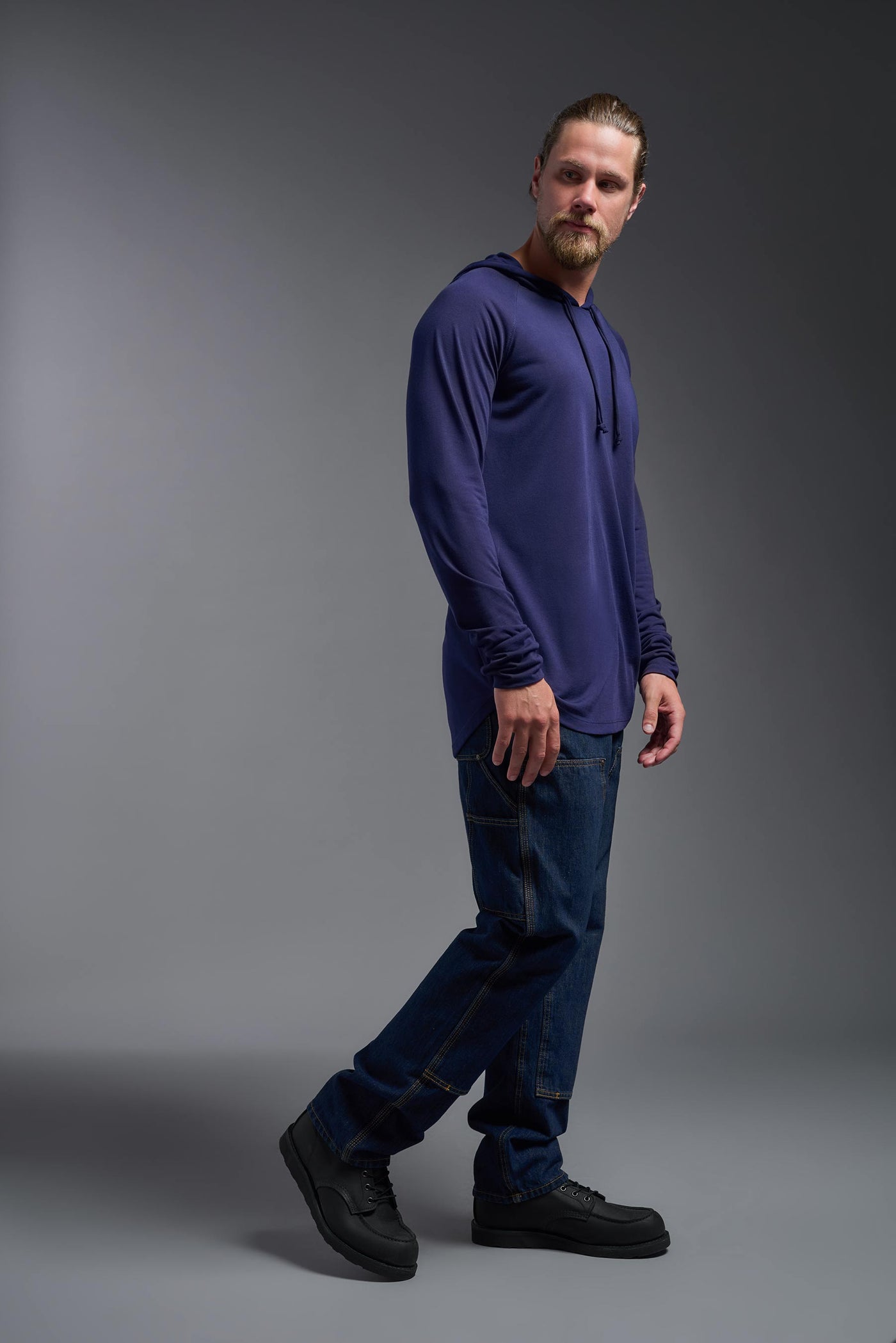 A blonde male model with a beard wearing the men's Siro Curvy Hoodie Shirt in the color navy, made out of 100% polyester, showing the relaxed fit from a side view. This hoodie shirt features a soft cotton feel, quick-dries, wicks moisture and is made in the USA. #color_navy