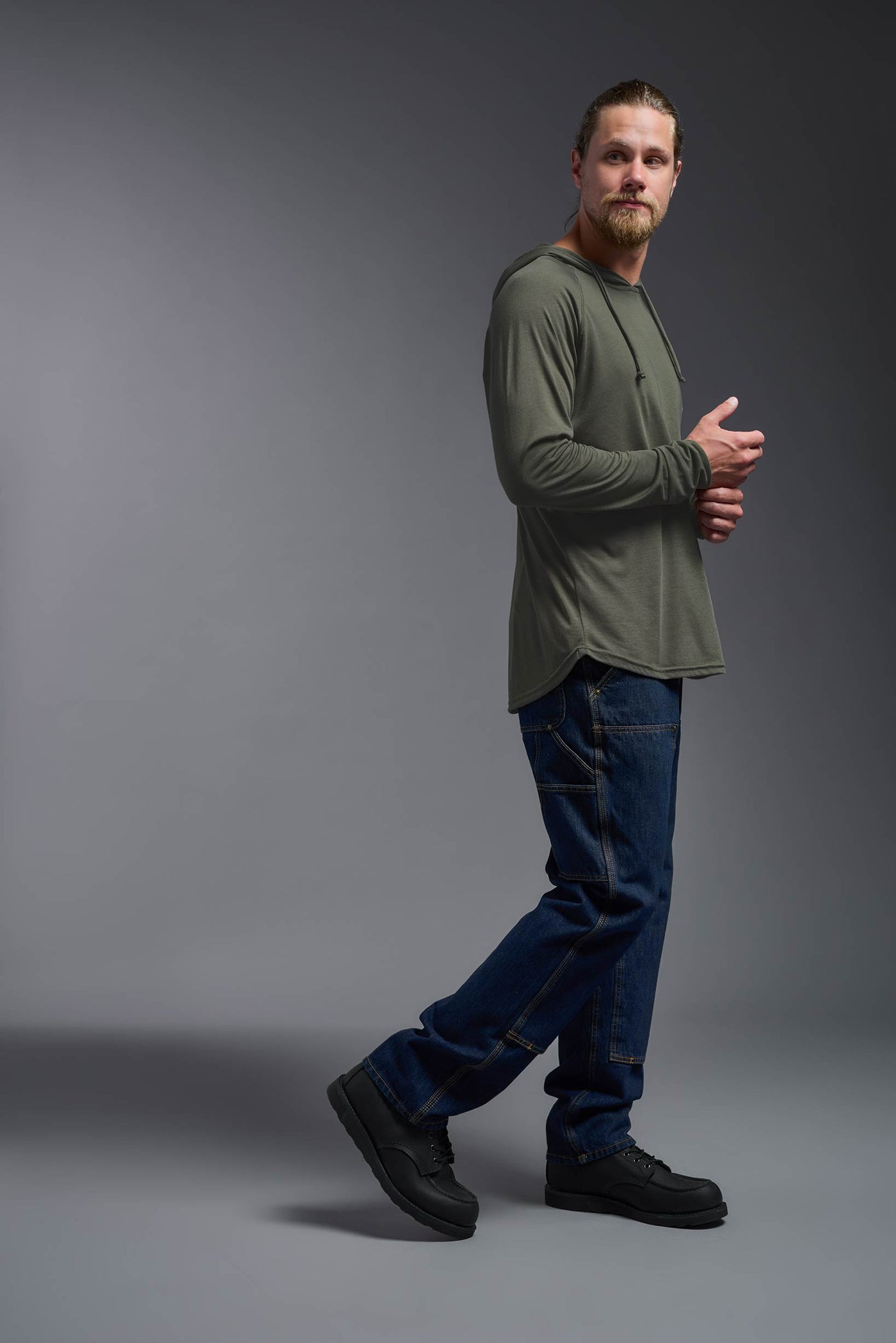 A blonde male model with a beard wearing the men's Siro Curvy Hoodie Shirt in the color olive, made out of 100% polyester, showing the relaxed fit from a side view. This hoodie shirt features a soft cotton feel, quick-dries, wicks moisture and is made in the USA. #color_olive