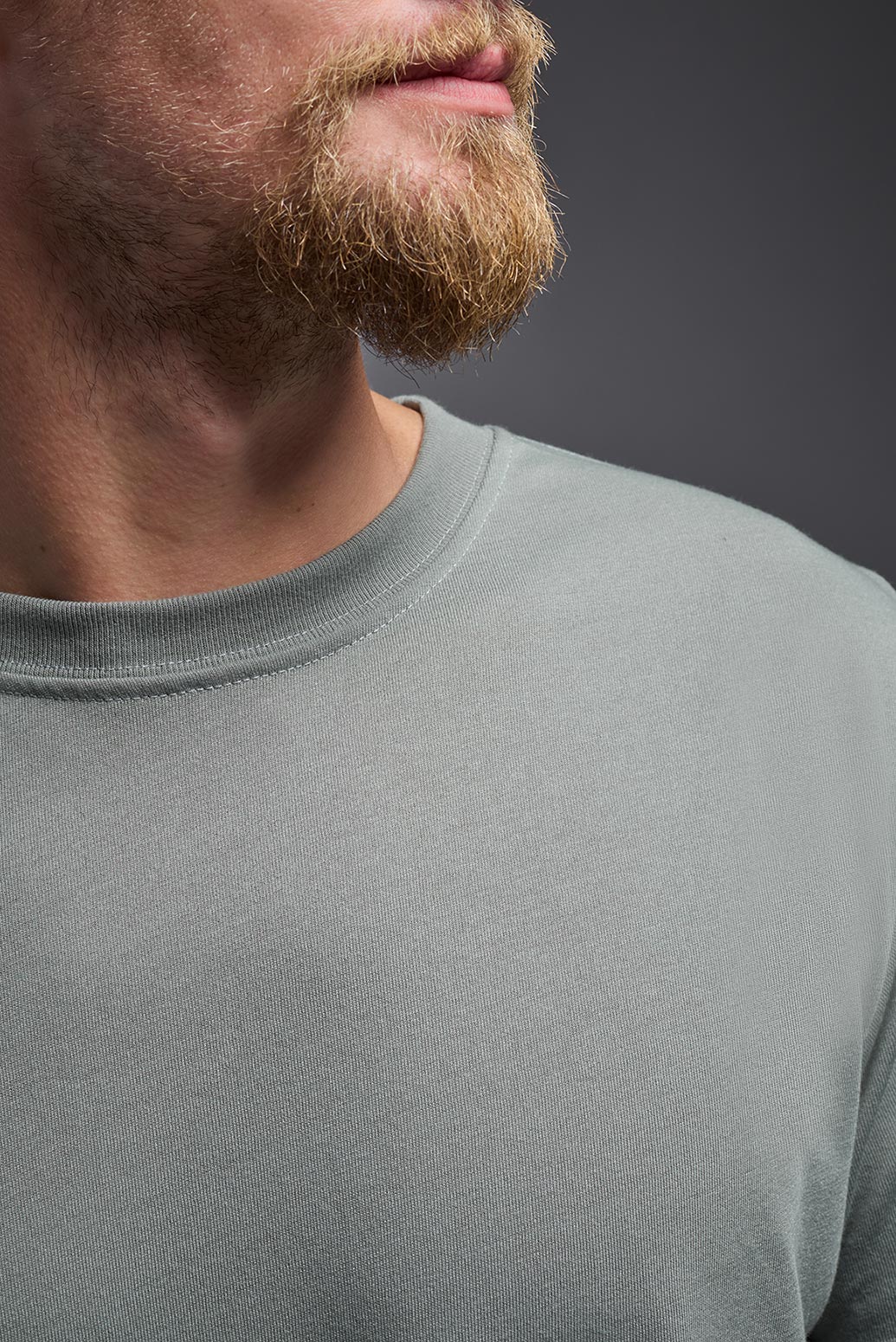 A male caucasian model with a beard wearing the men's DuroSoft Outdoor Tee in the color army gray on a gray background, made out of 100% filament polyester that feels like soft cotton, showing the modern fit from a close-up collar view. This shirt is anti-snagging, quick-drying and moisture-wicking and is made in the USA. #color_army-gray